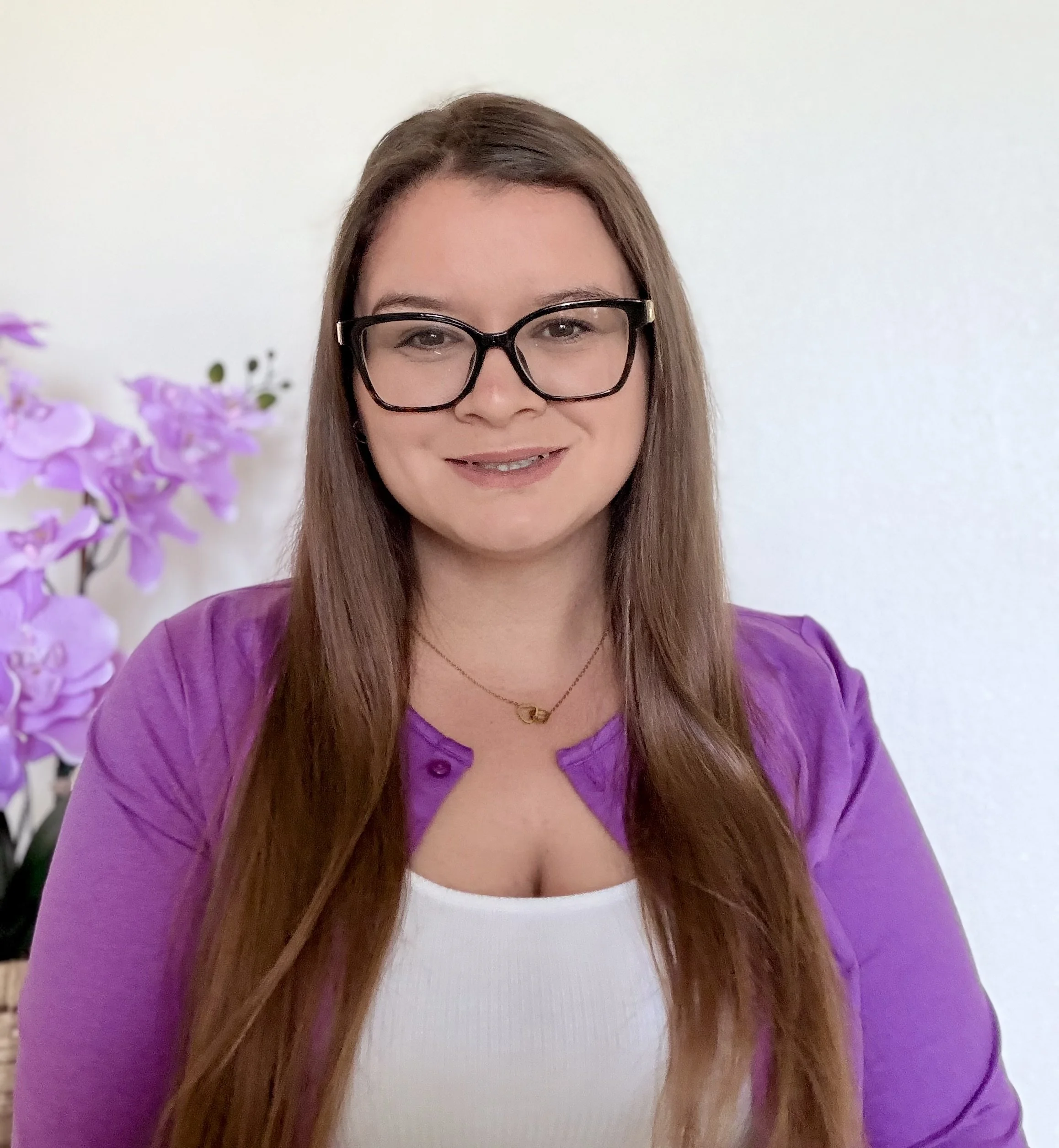 Photo of a woman with long brown hair, wearing large black glasses, a white top, and a purple jacket, standing indoors with a plain white wall and purple flowers in the background.