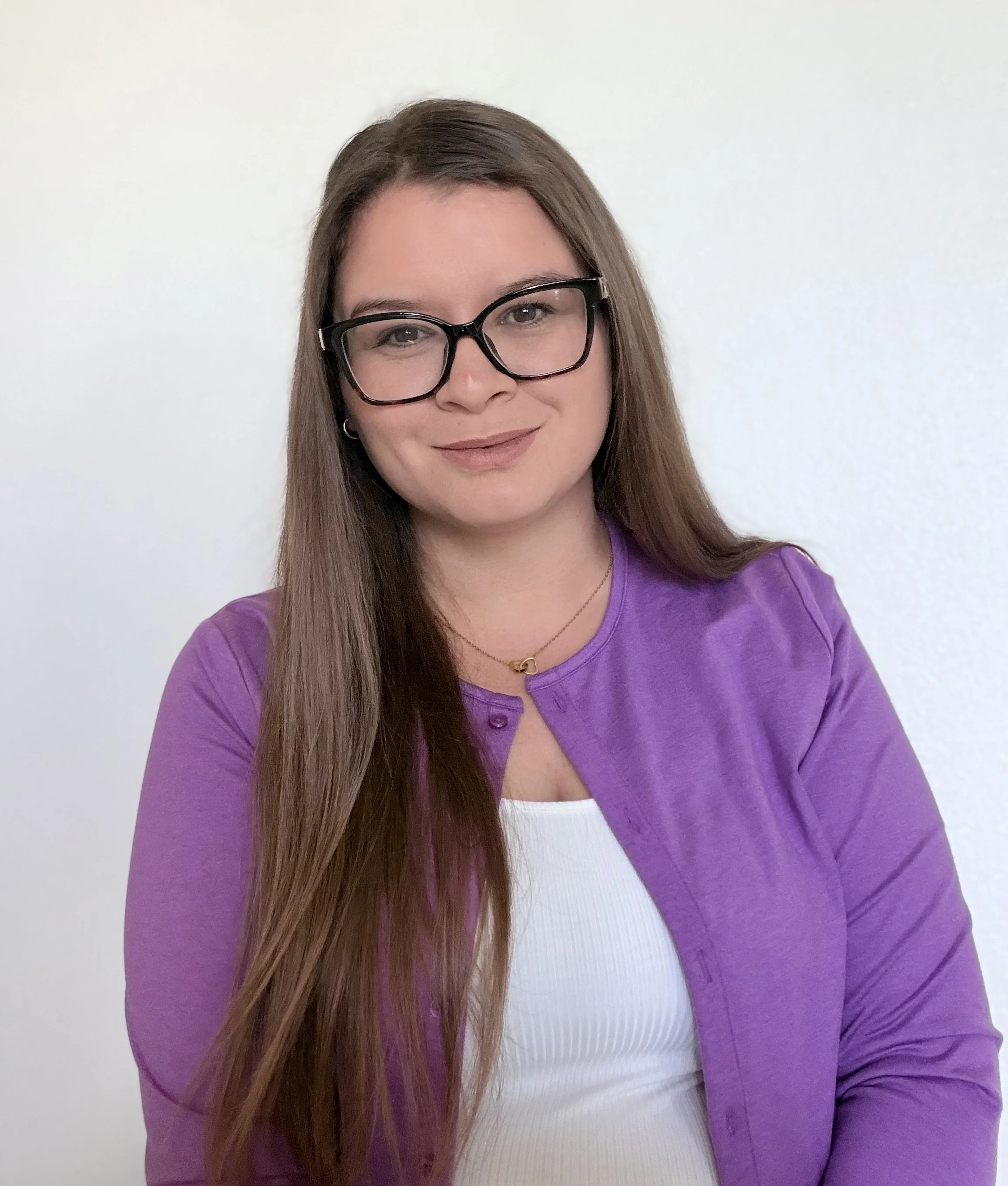 A woman with long brown hair, wearing black glasses, a purple shirt, a white top underneath, and a gold necklace, standing against a plain light-colored wall.