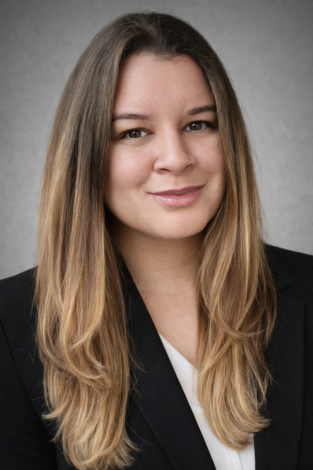 A professional woman with long light brown hair, wearing a black blazer and white top, smiling slightly, against a gray background.