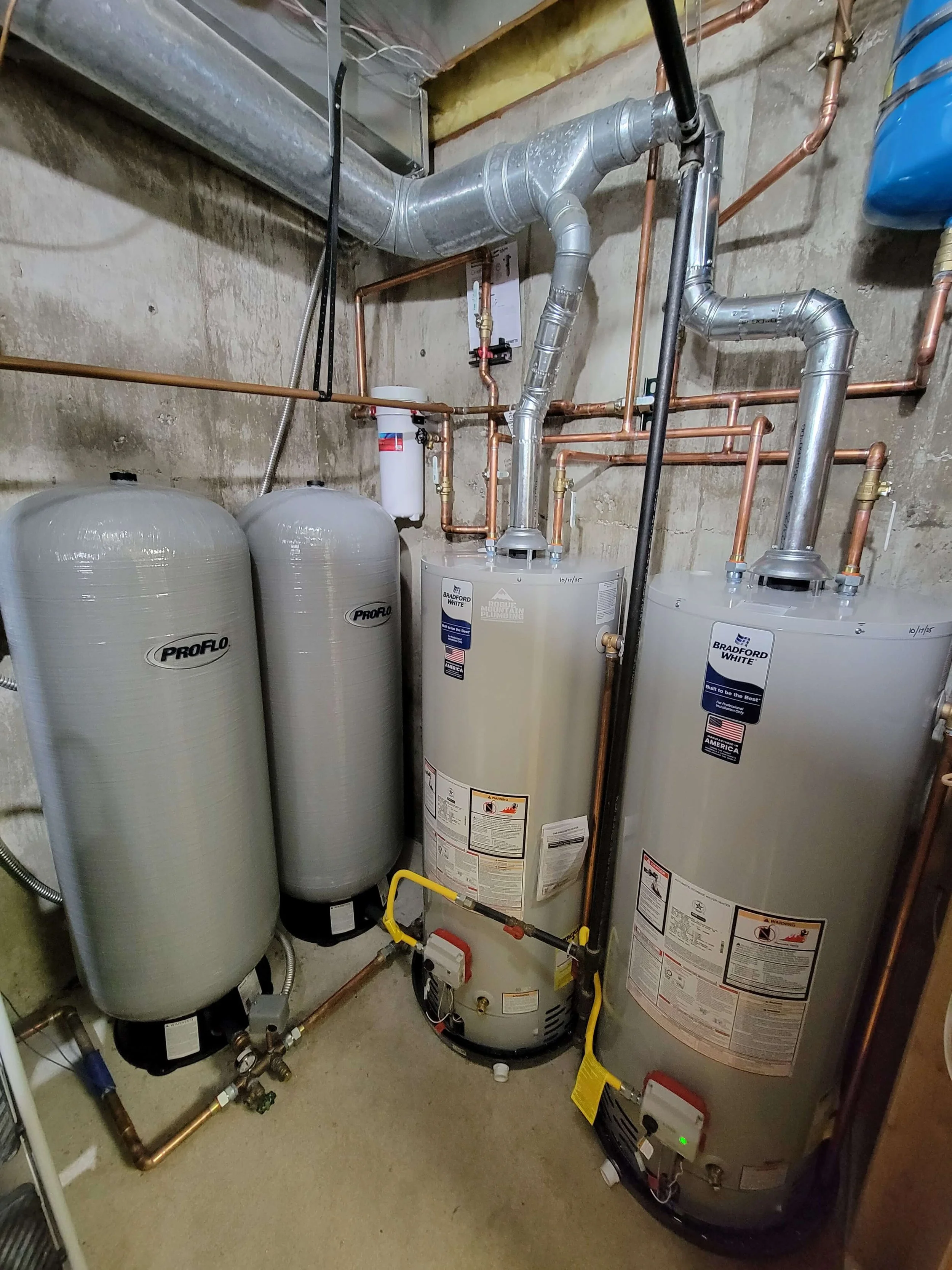 Three water heaters and water filtration system in a utility room with copper and metal piping.
