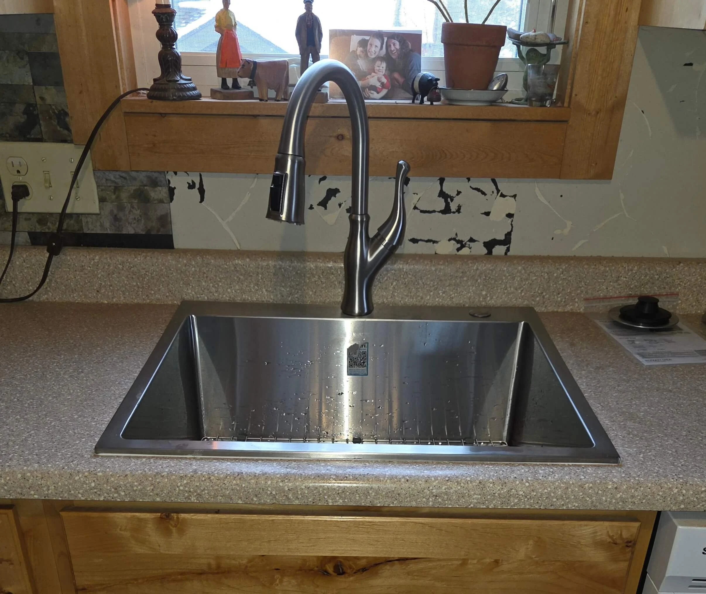 Silver kitchen sink with water droplets and a modern faucet, set in a speckled beige countertop with a wooden cabinet below, and a window with decorative items on the windowsill in the background.