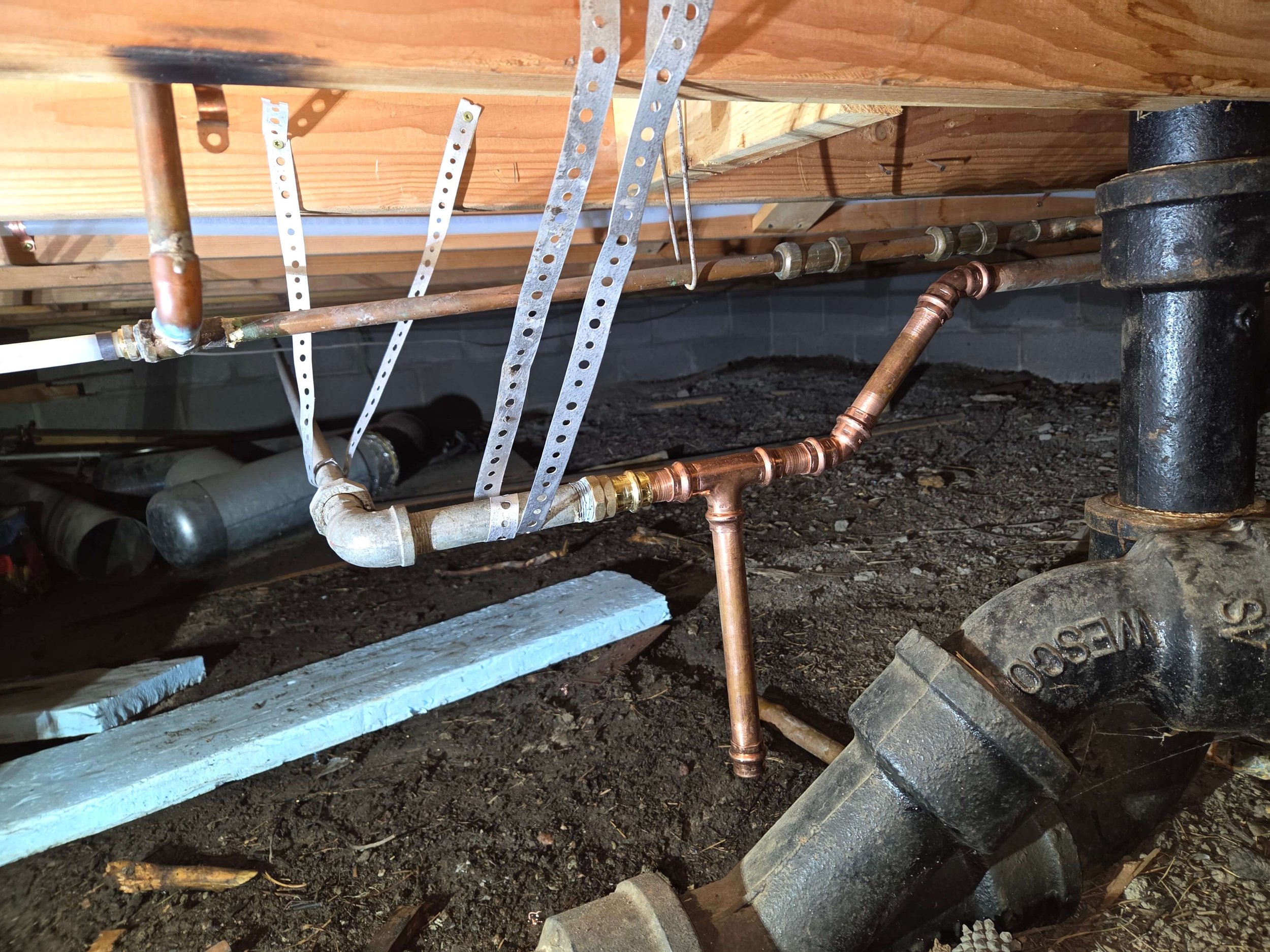 Copper and PVC plumbing pipes under a house with metal straps for support, connected to a large black cast iron drainage pipe.