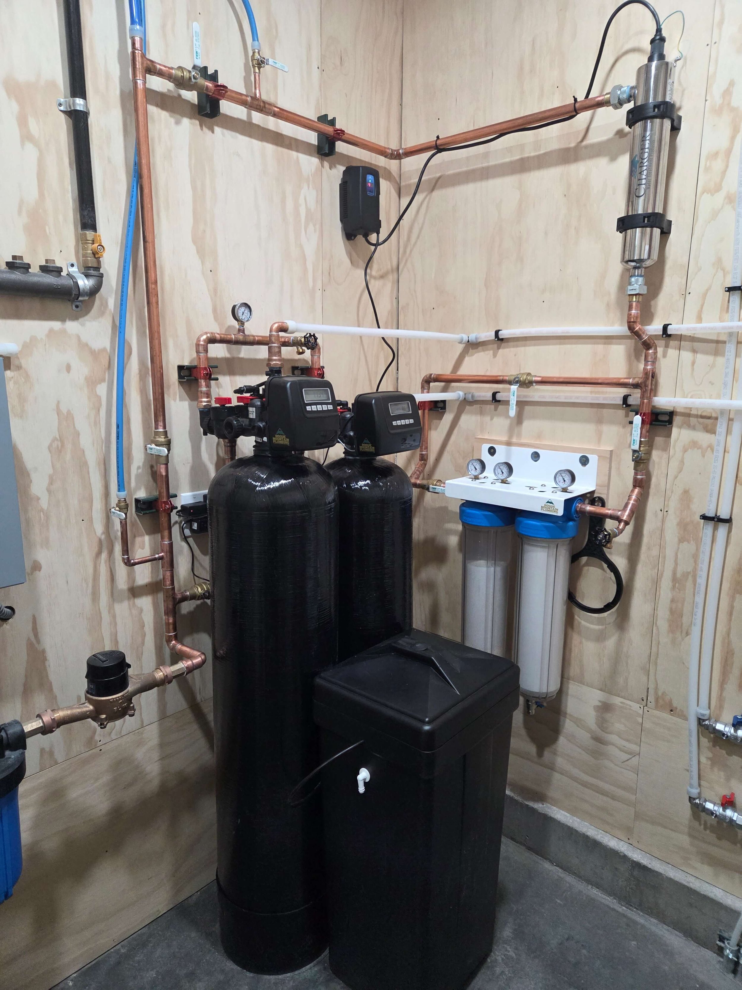 Water treatment system with black tanks, white filter housings, and copper plumbing, installed on a wooden wall in a utility room.