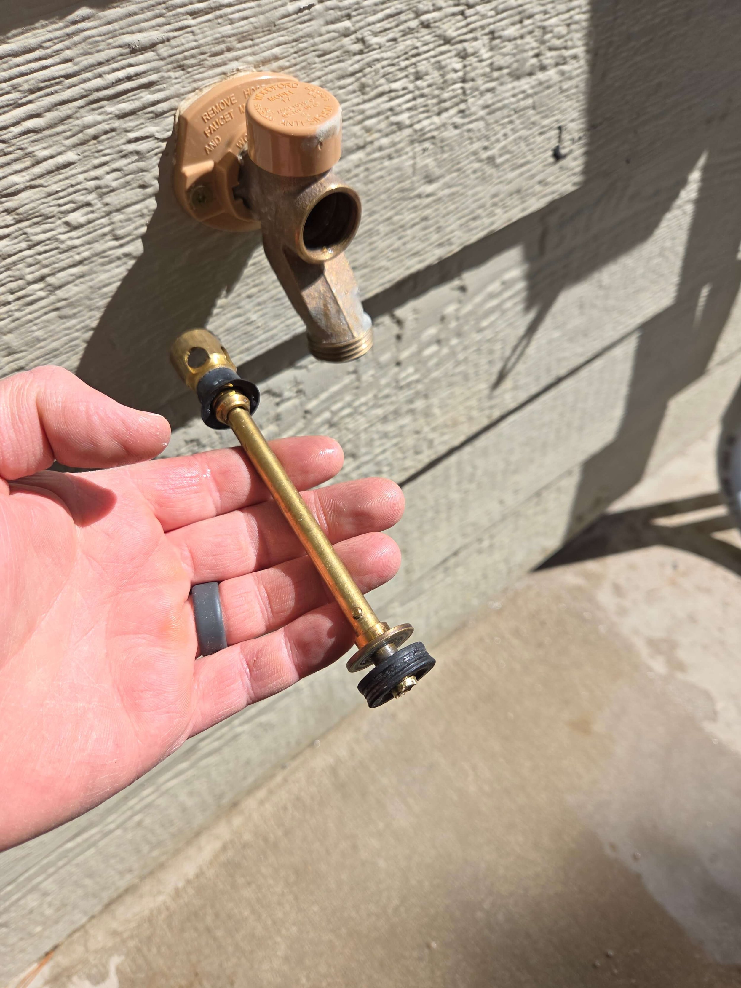Outdoor hose bib with removed faucet stem held in a hand, against a wooden wall.