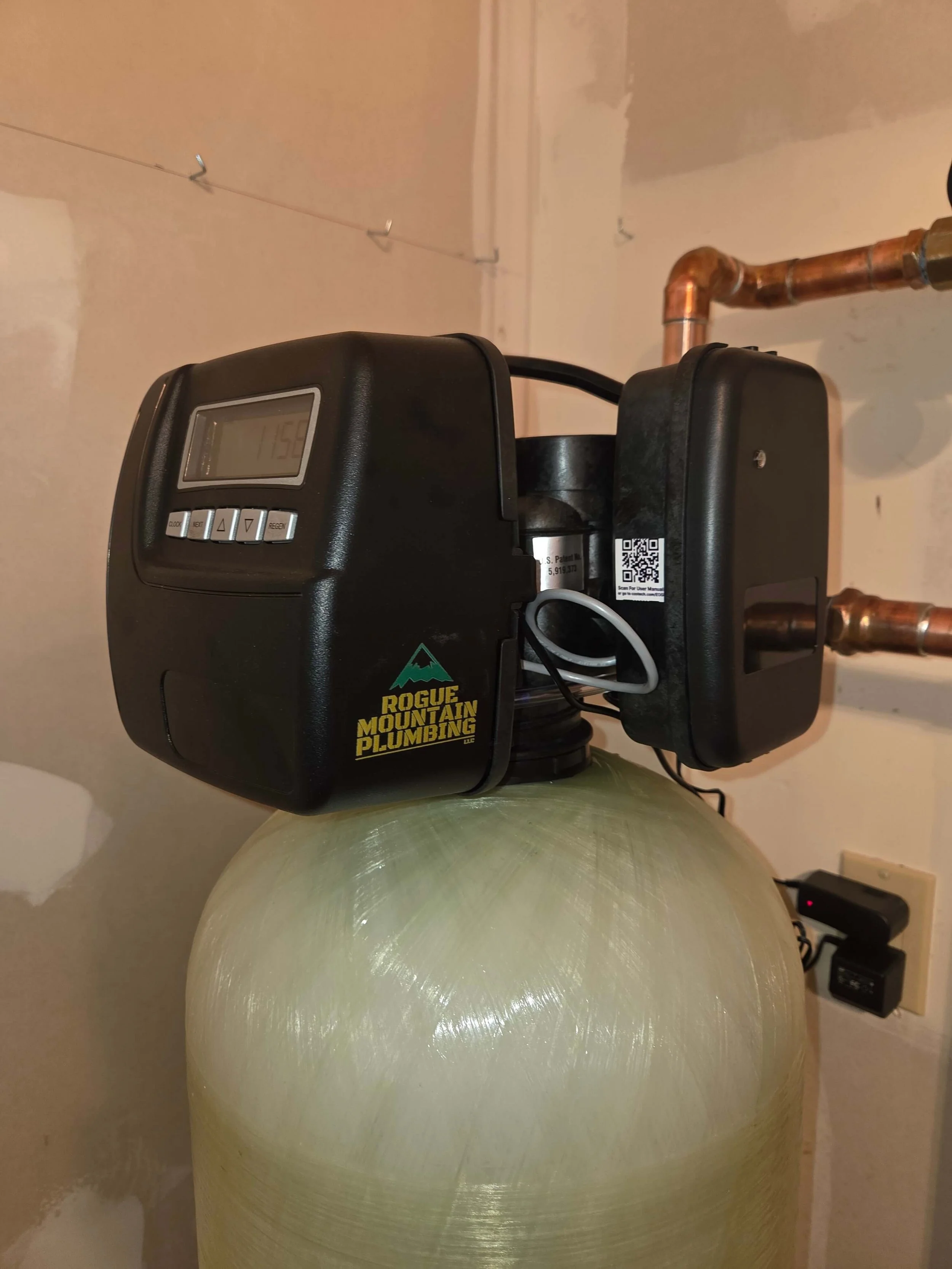 A water softener unit with a digital control panel on top, placed on a large, rounded tank, with copper pipes connected to it.