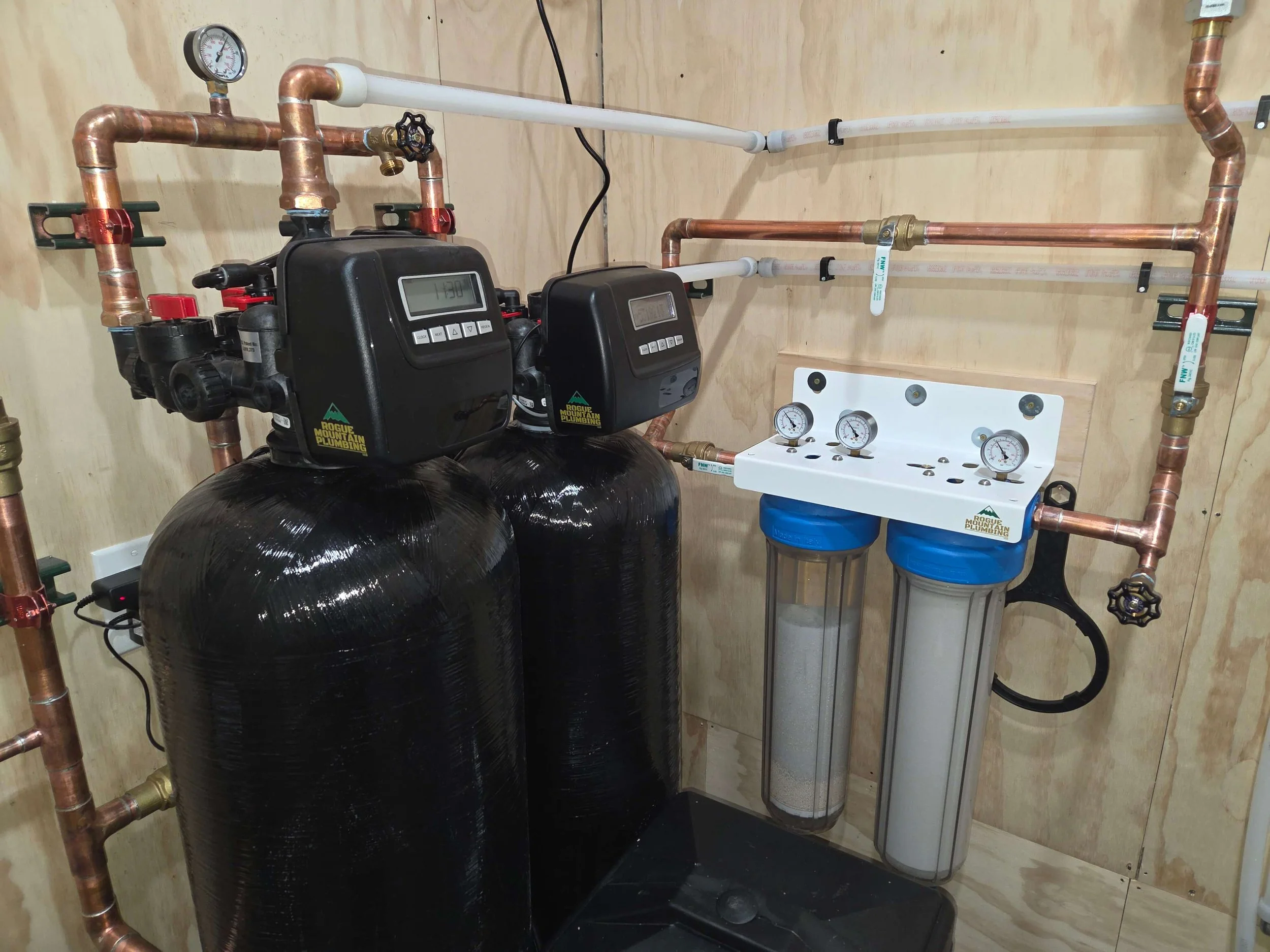 An indoor water filtration and treatment system with two black tanks, water pressure gauges, and white pipes and valves.