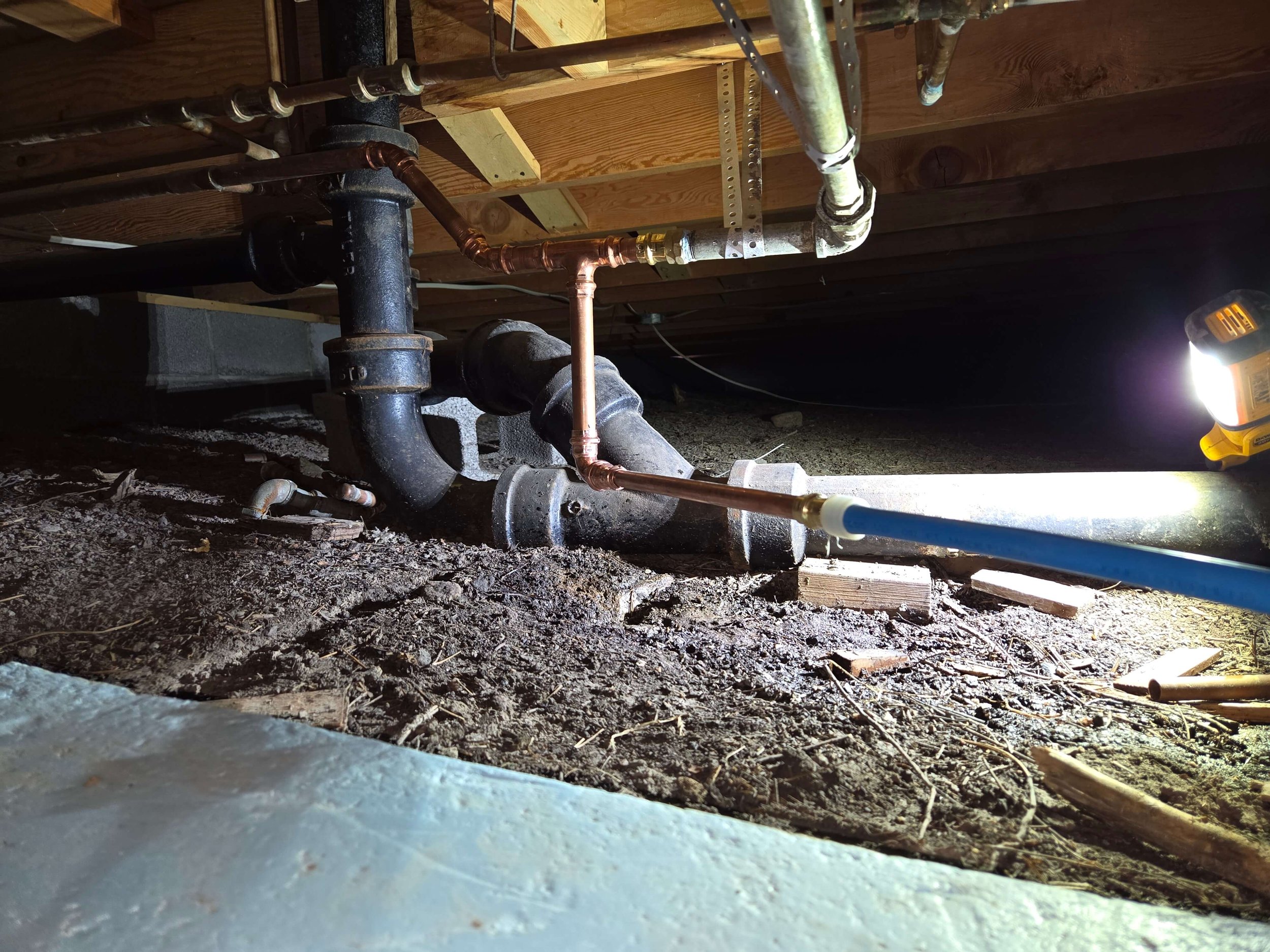 View of plumbing pipes under a house, including black cast iron drainage and copper pipes, with a flashlight illuminating the area.