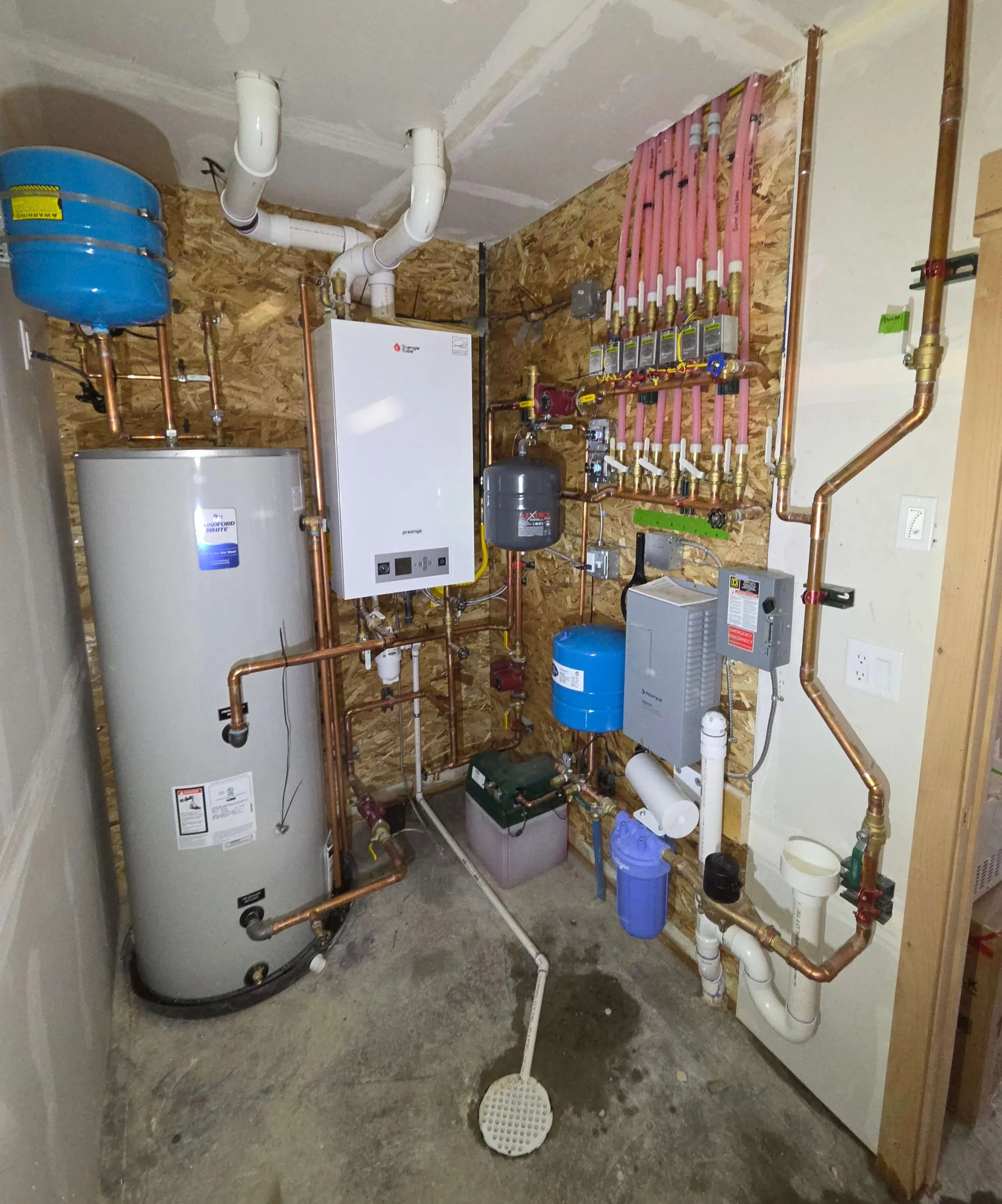 A utility room with plumbing, a water heater, and a boiler system installed on a plywood wall.