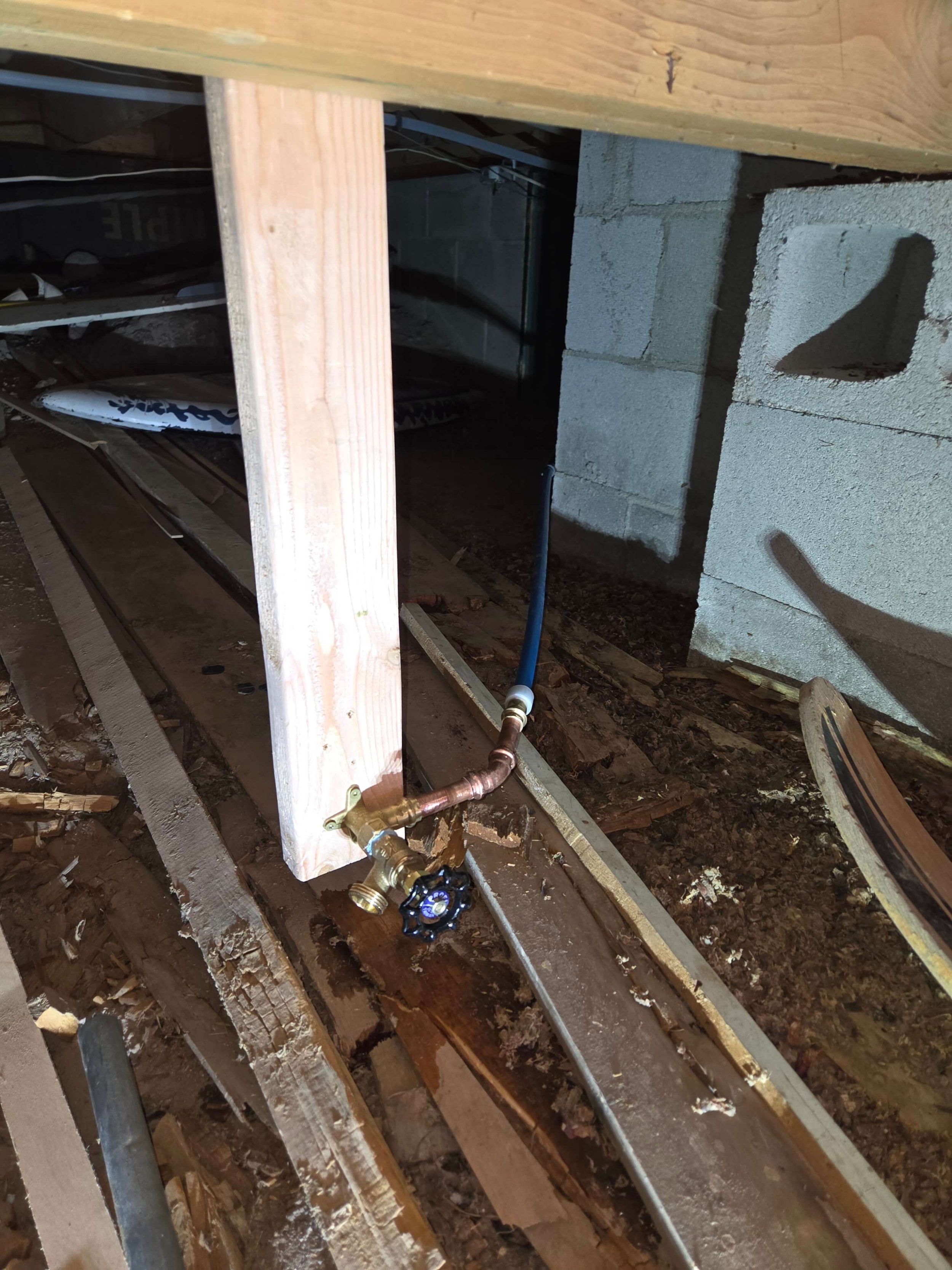 Indoor plumbing setup with a water valve attached to a wooden post, blue hose connected to a brass fitting, and concrete blocks in the background.