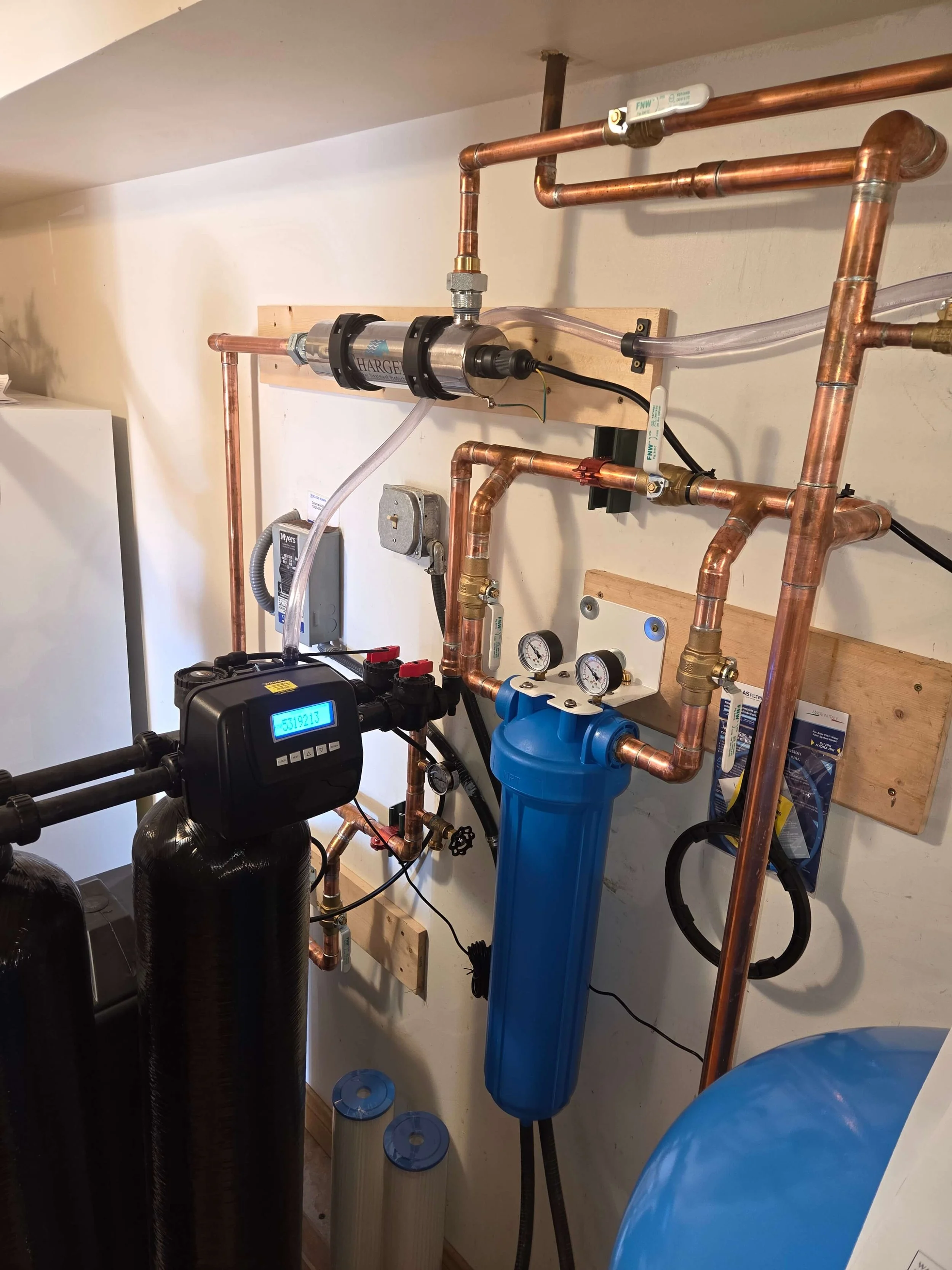 Water filtration system with copper pipes, a blue filter housing, and gauges mounted on a wall.