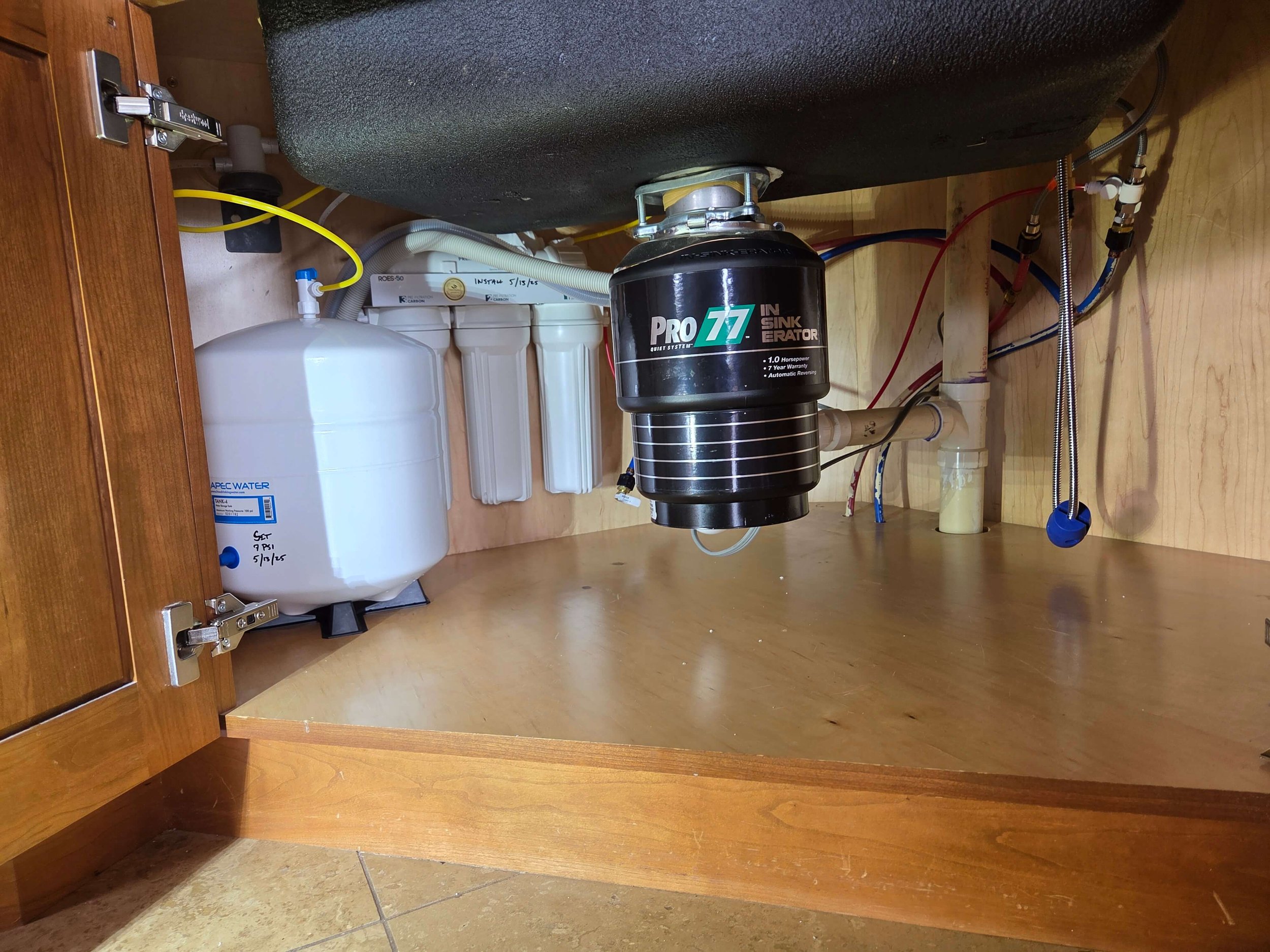Under-sink view in a kitchen showing a garbage disposal unit, water filtration system, and pipes. A Pro77 InSinkErator is mounted under the sink and a white tank labeled ApecWater is visible, alongside various plumbing connections.
