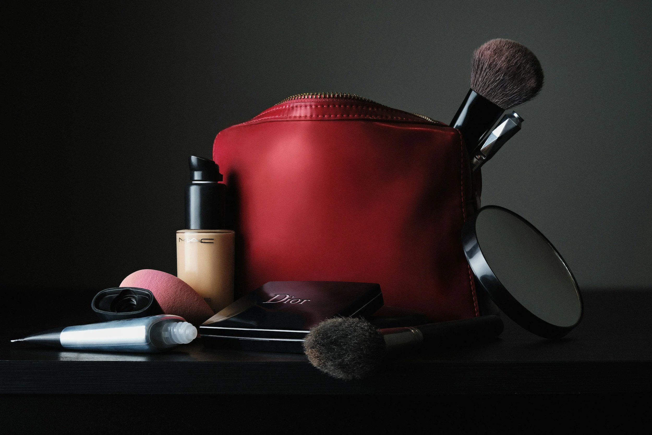 Beauty products and accessories arranged on a black surface, including a red makeup bag with a zipper, makeup brushes, foundation bottle, sponge, and an eyeshadow palette, with a circular mirror in the background.