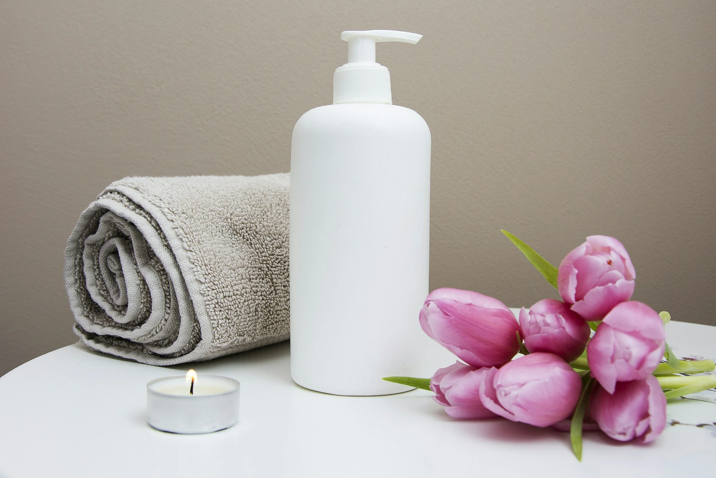 A spa or bathroom setting with a rolled grey towel, a white pump soap dispenser, a small lit tea light candle, and a bunch of pink peonies on a white surface with a beige wall background.