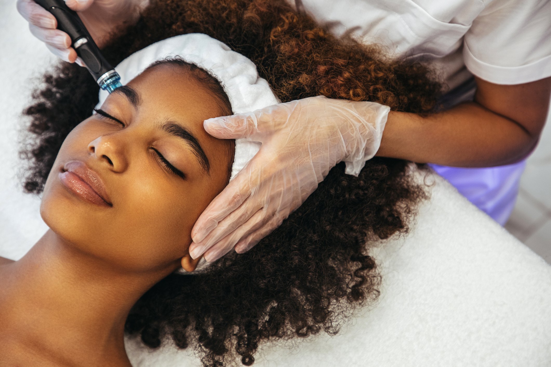 A woman lying down with her eyes closed, receiving a facial treatment from a professional wearing gloves. The professional is using a handheld device on the woman's forehead with her long, curly hair surrounding her head.