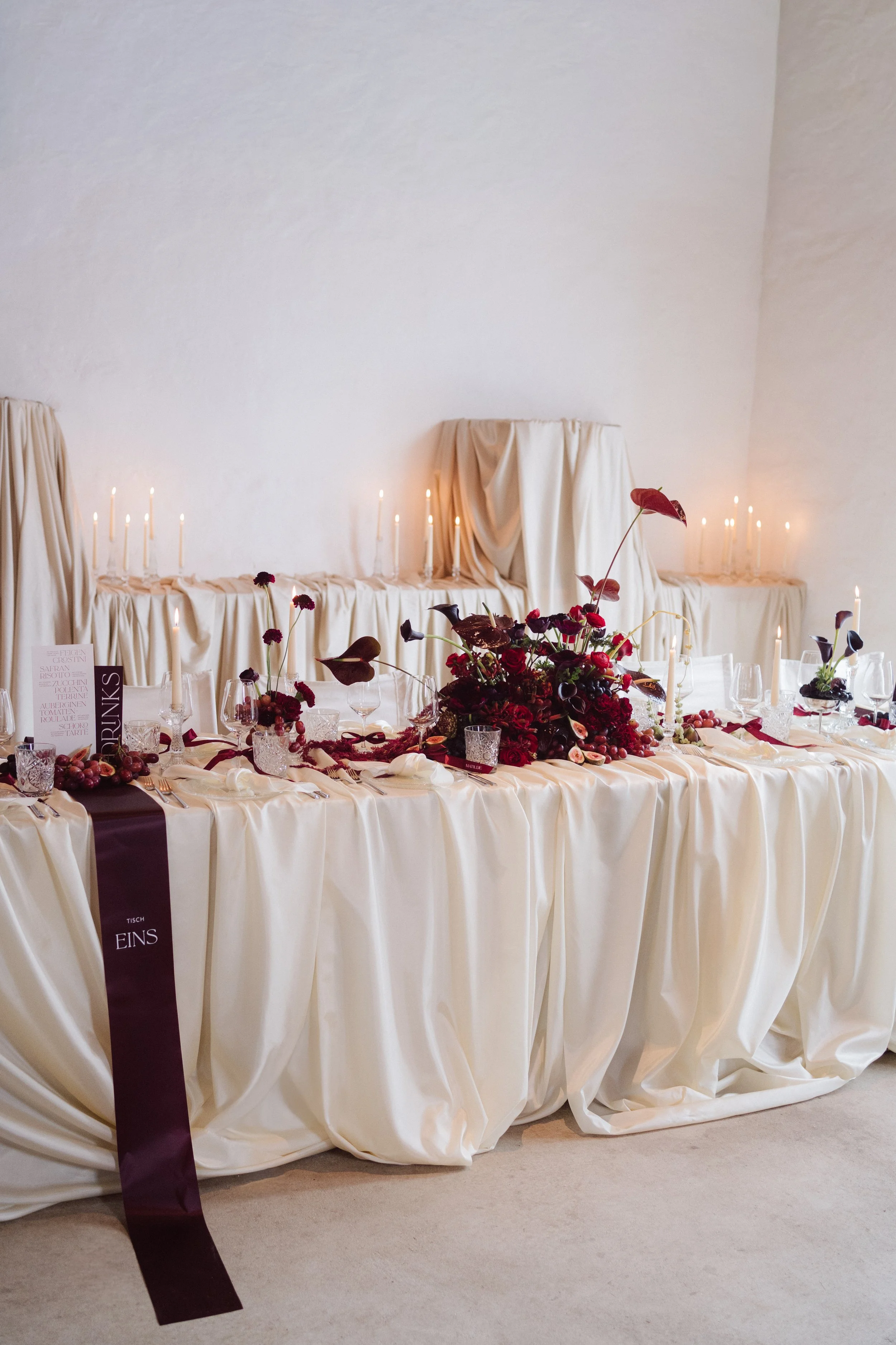 Brauttafel mit Kerzen und weinroten Blumen vor der Stoffwand