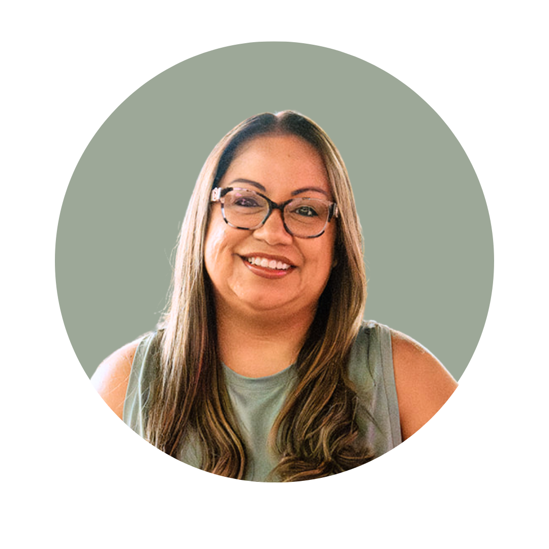Smiling person wearing clear glasses with red lipstick, brown hair, and a dark top, against a grey background.