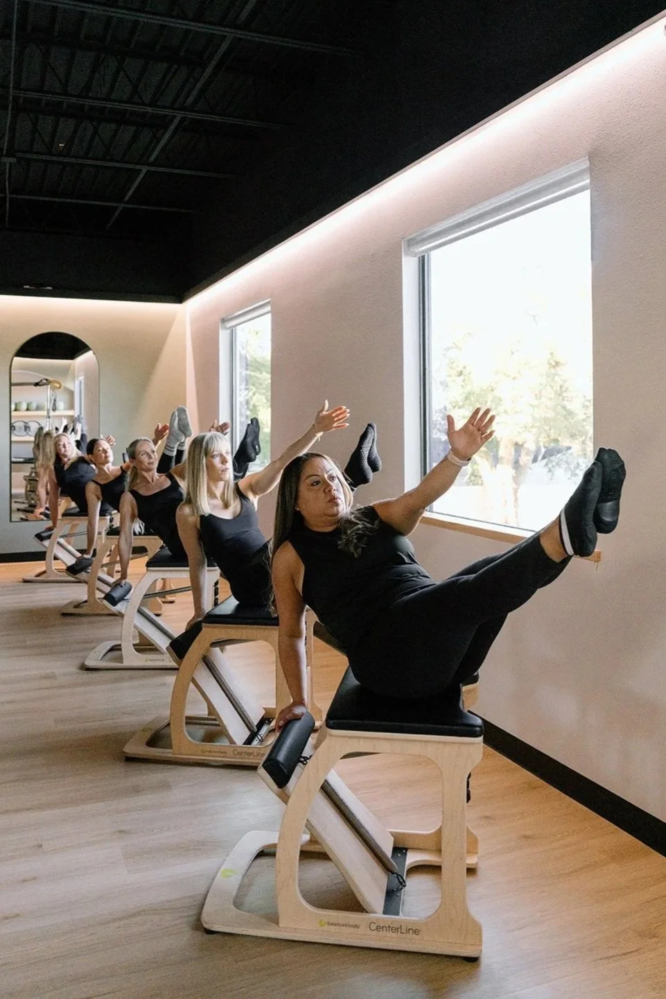 A group of three women practicing Pilates on reformer machines, each holding a Pilates ring, in a studio with wooden floors and a quote on the wall.