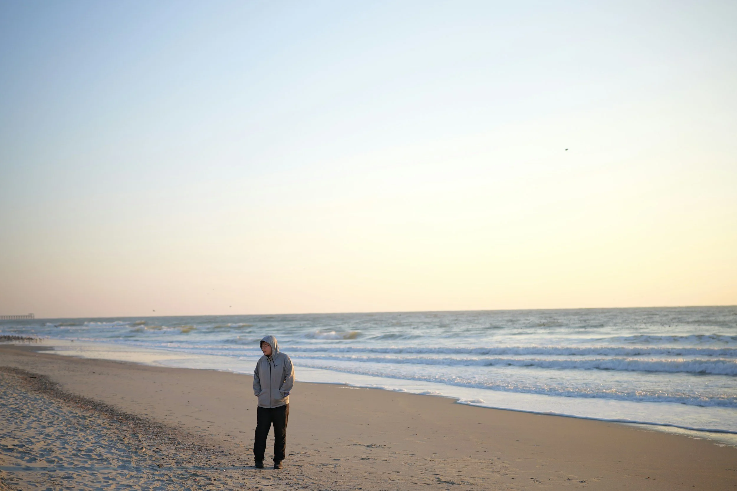 Person in a gray hoodie and black pants walking alone on a sandy beach at sunset, with waves in the background.
