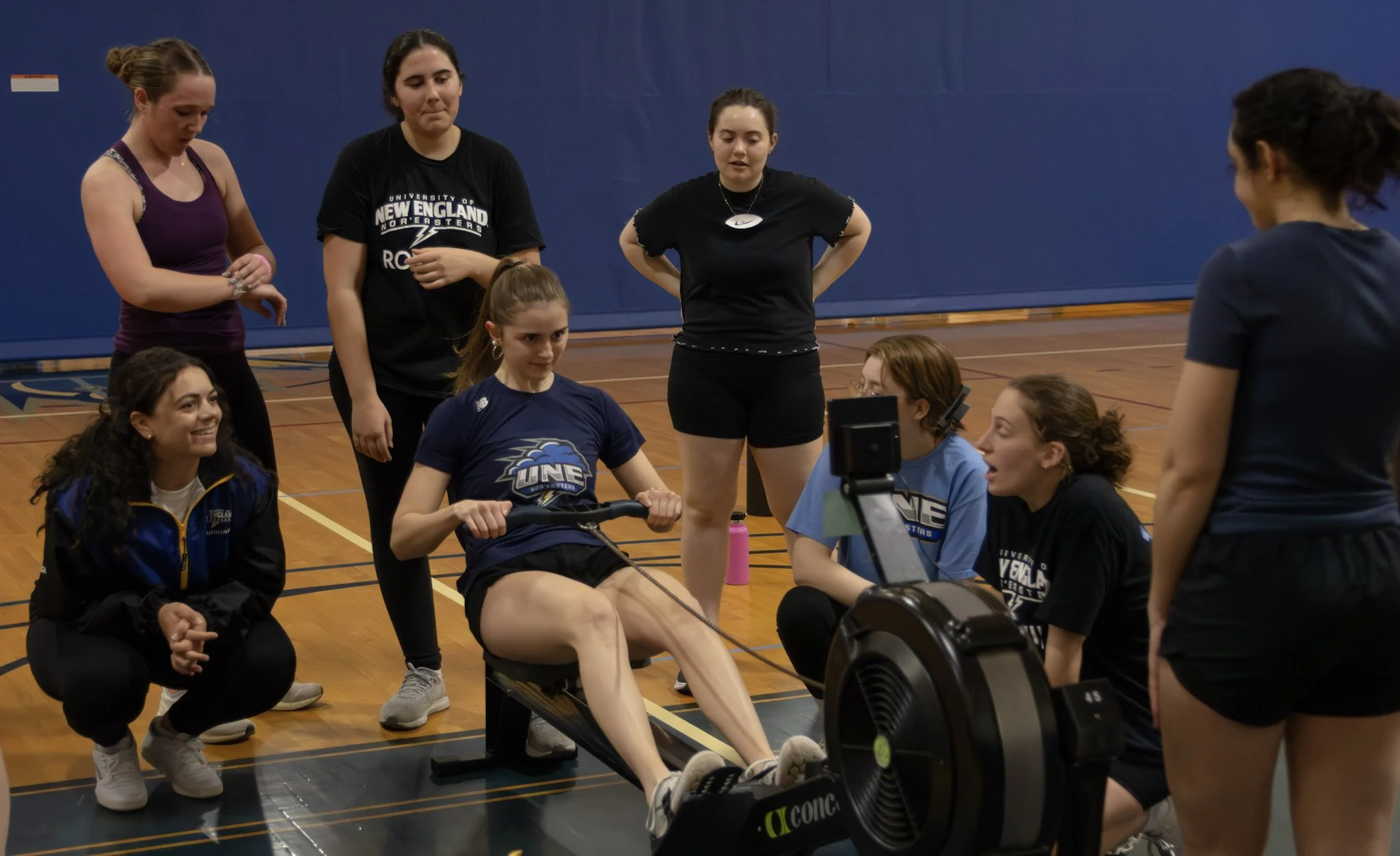 A group of young adult rowers are gathered around another rower on an erg. A coach is crouched down smiling and supporting