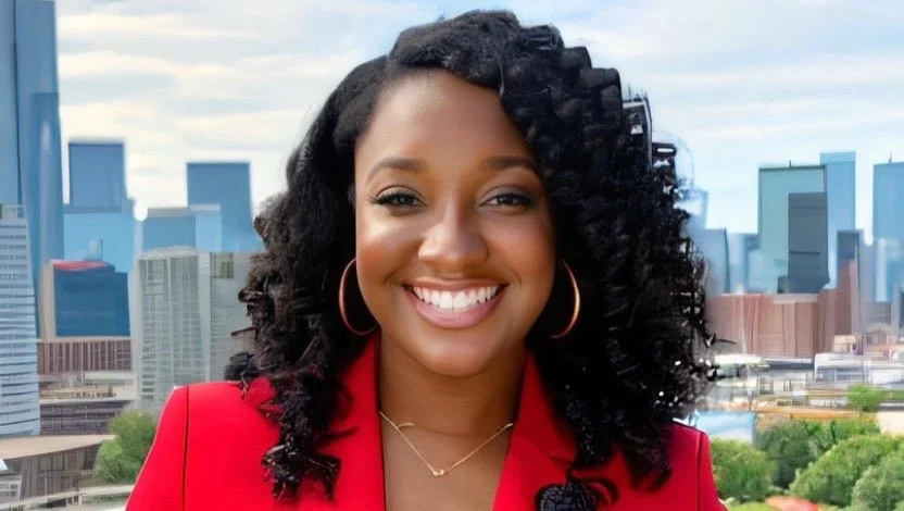 A woman with curly black hair and hoop earrings smiling outdoors in front of a city skyline.