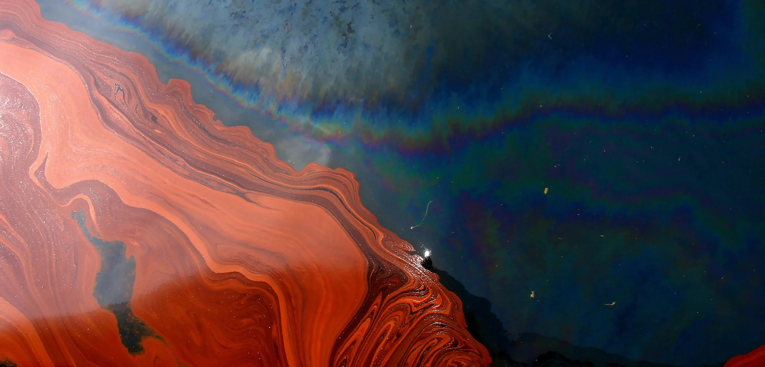 Aerial view of colorful mineral deposits and water, showing swirling red, orange, and pink patterns with dark patches and a rainbow-colored streak across the water.