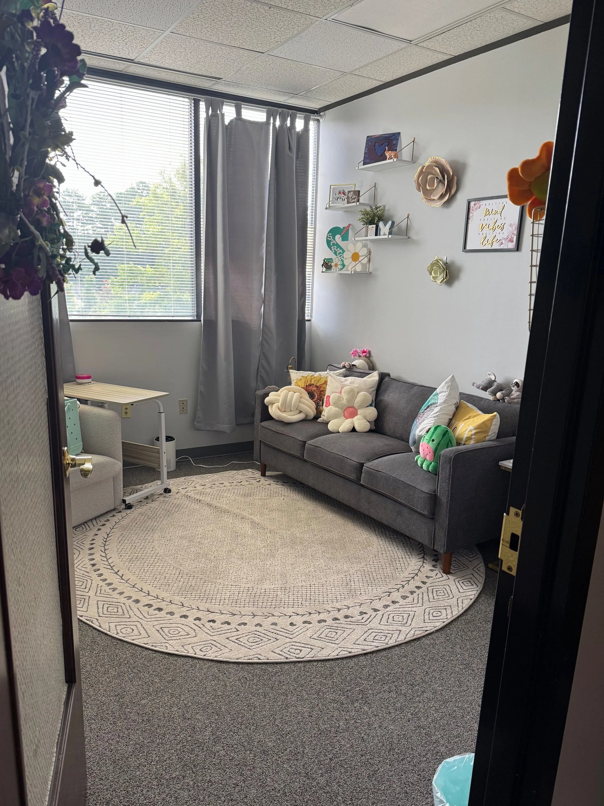A cozy office or lounge with a gray sofa, decorative pillows, wall shelves with plants and art, a window with blinds and curtains, and a round woven rug on gray carpeted floor.