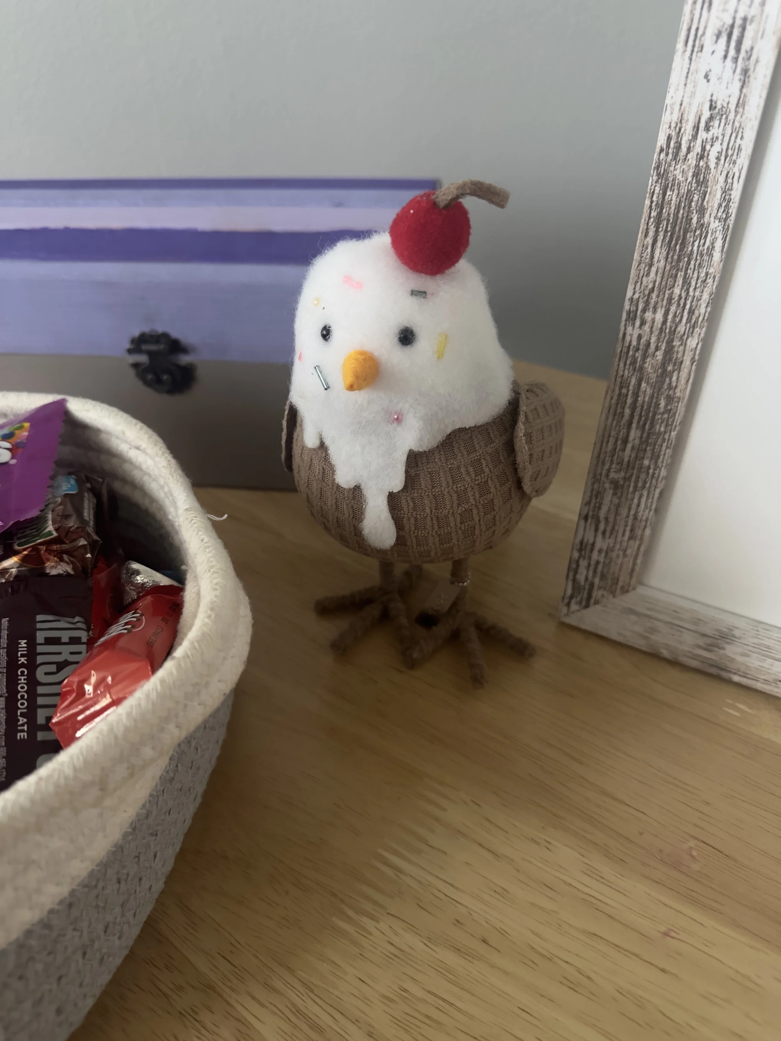 A whimsical chicken figurine with a sock-wool body, decorated with cake-like white frosting and sprinkles on its head, a cherry on top, and an orange beak, standing on a wooden surface next to a basket of snack bags and a picture frame.