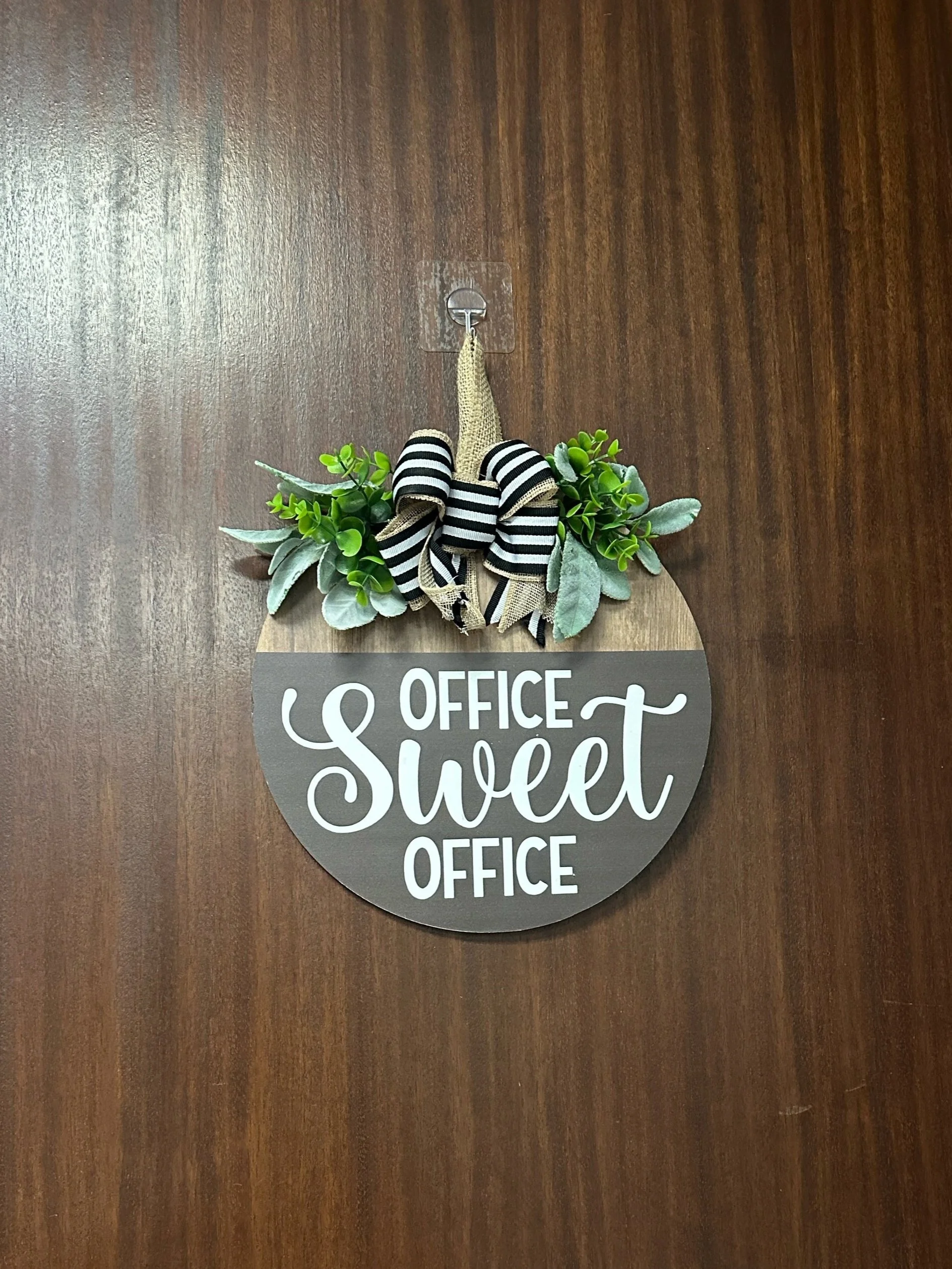 Decorative circular sign on door with the words 'Office Sweet Office' surrounded by artificial greenery and a black and white striped bow.