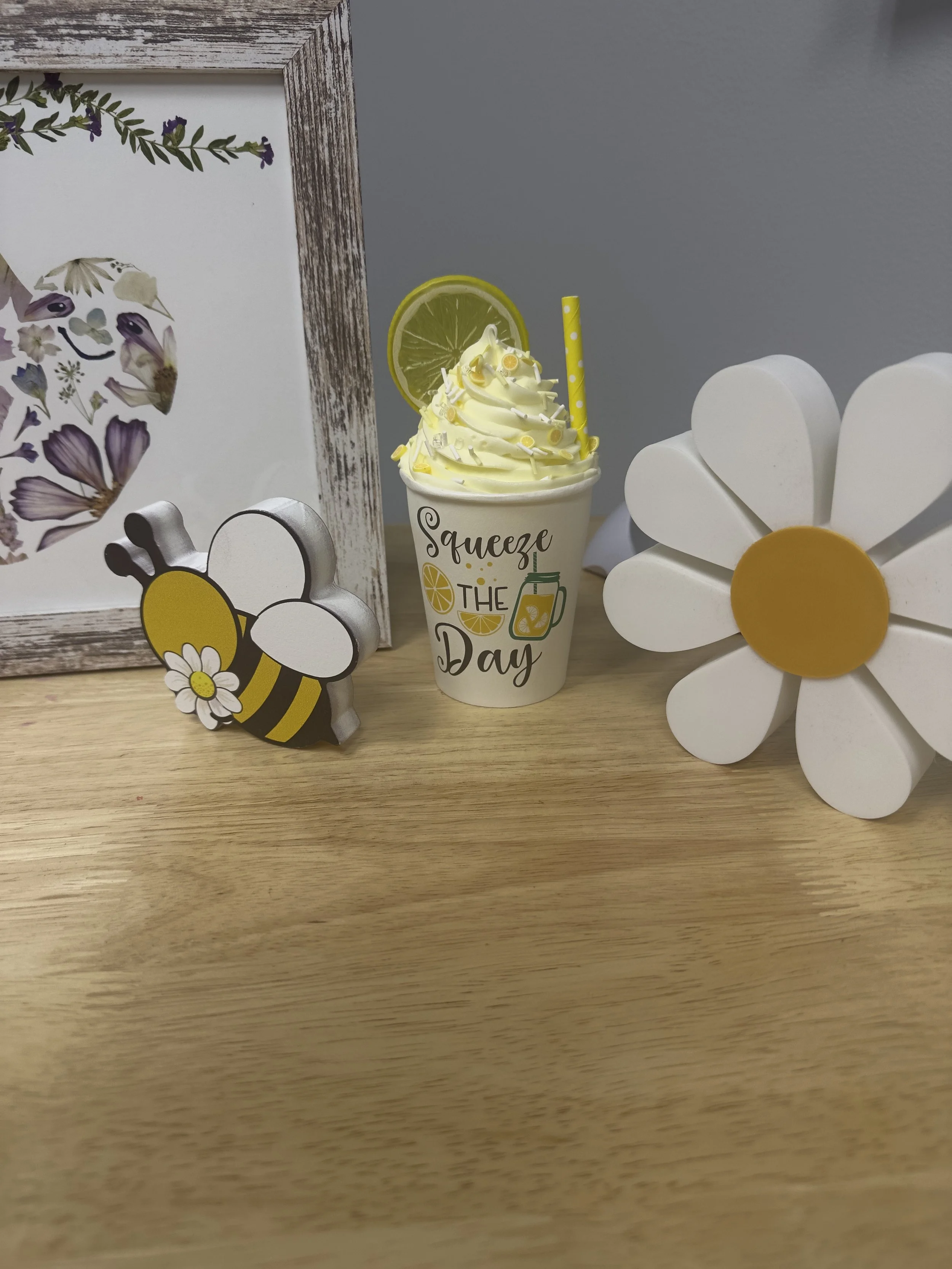 Decorative desk scene featuring a framed botanical print, a black and yellow bee figurine with a white flower, a white flower decoration with a yellow center, and a yellow lemon-themed beverage with whipped cream, lemon slice, and a yellow polka dot straw, with a gray background.