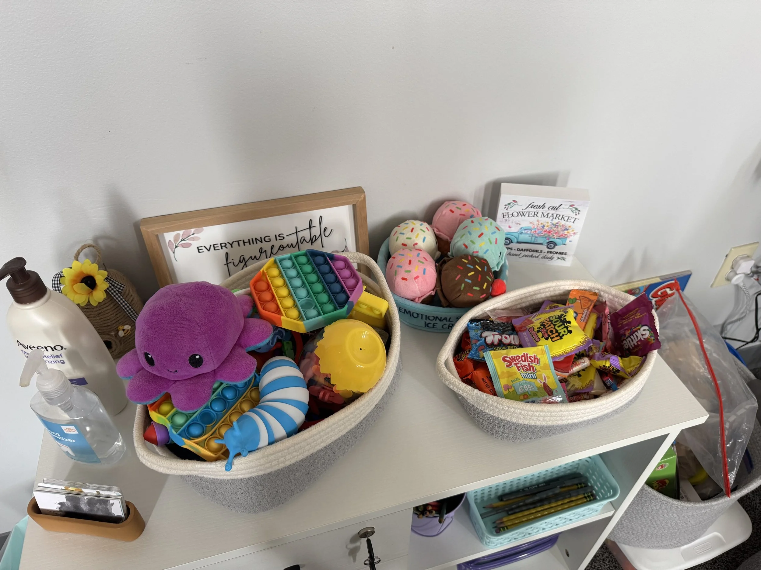 Baskets filled with children's toys, plush toys, colorful squishy toys, and wrapped candies on a white table against a white wall with a hand sanitizer, framed quote, and decorative items.