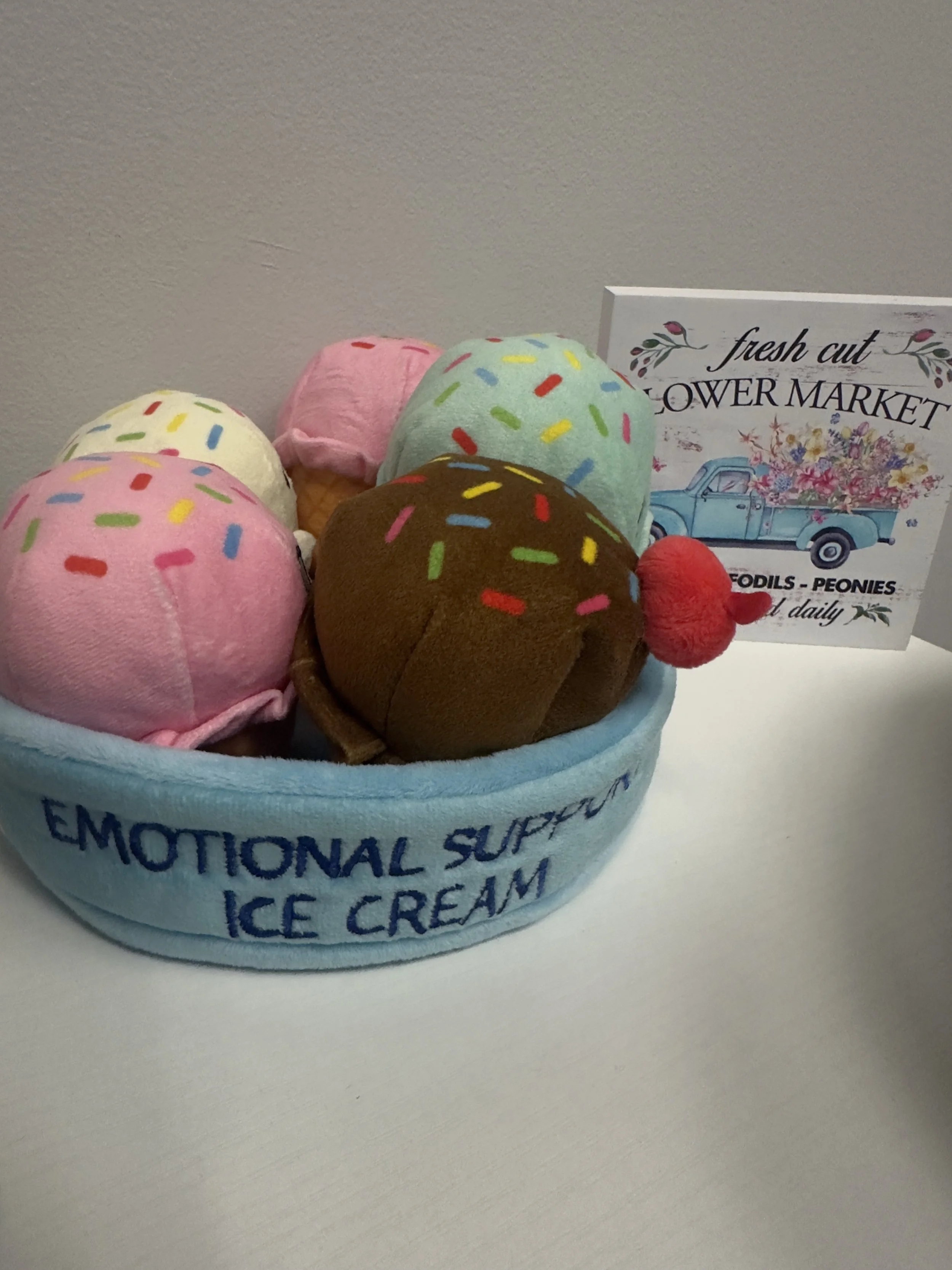 Felt plush ice cream cones in a blue bowl labeled "Emotional Support Ice Cream," with a sign behind them reading "Fresh Cut Flower Market" and featuring a vintage truck with flower arrangements.