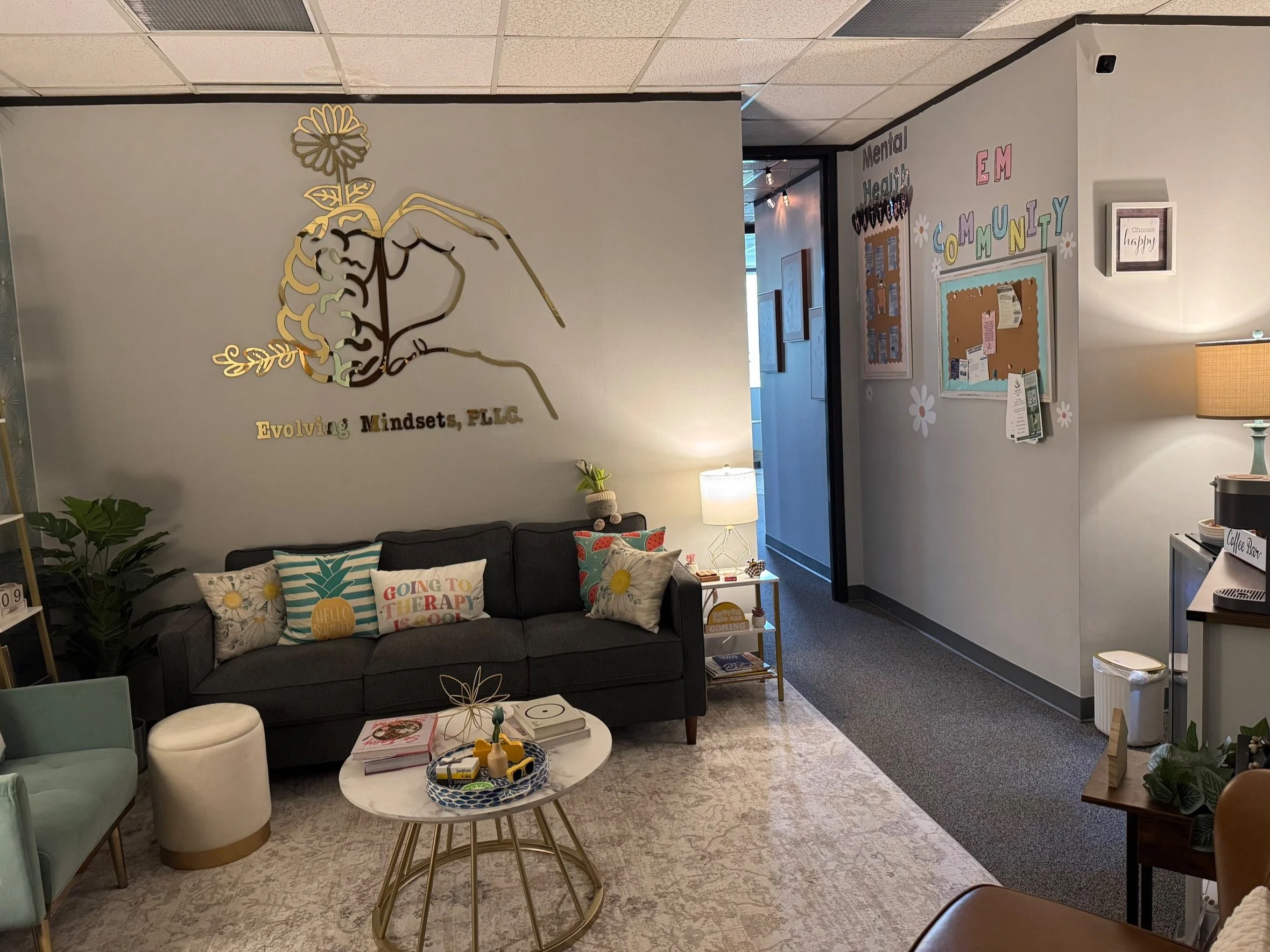 Waiting room with dark sofa, decorative pillows, side table with lamp, corner with large plant, decorative wall art reading "Evolving Mindsets, PLLC," another wall with bulletin boards, and a coffee station.