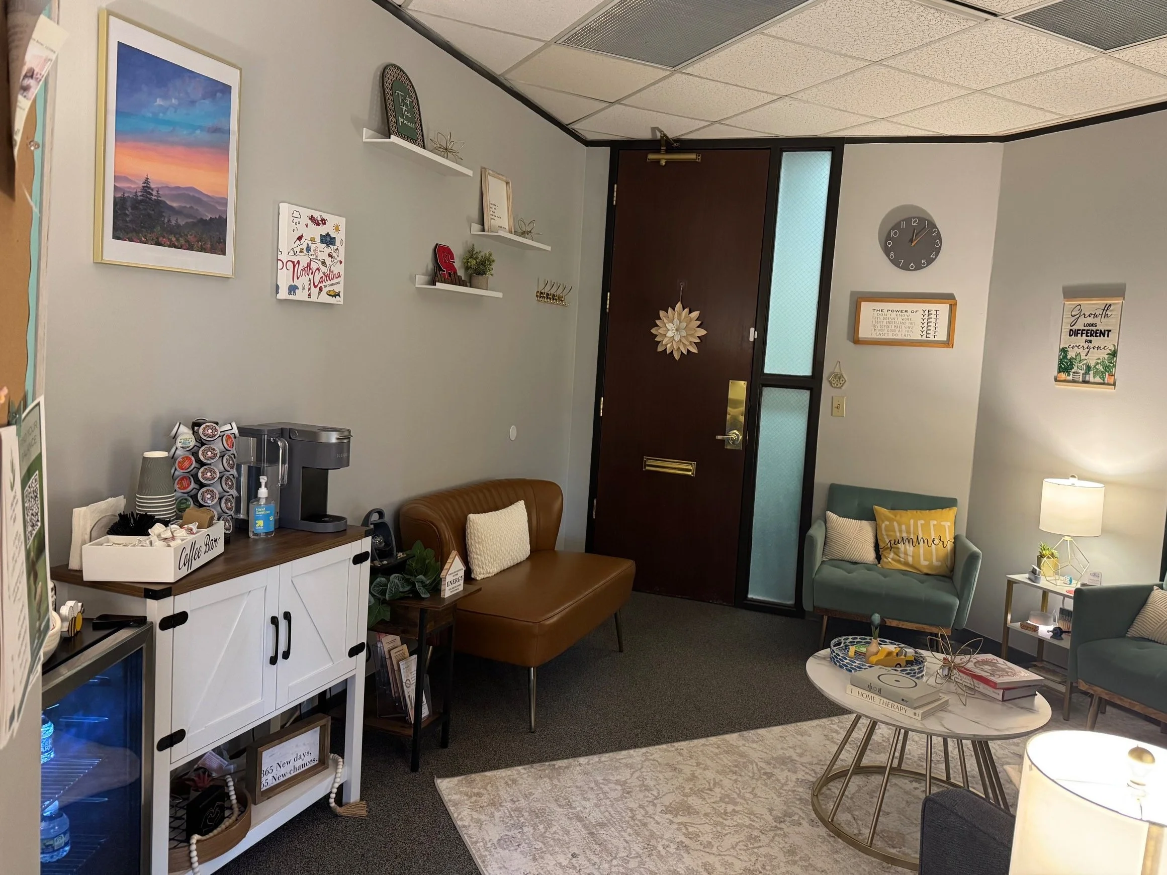 A cozy waiting room with a white and brown coffee station, two mint green chairs with pillows, a white table with decorative objects, and walls decorated with framed art, shelves, and a clock.