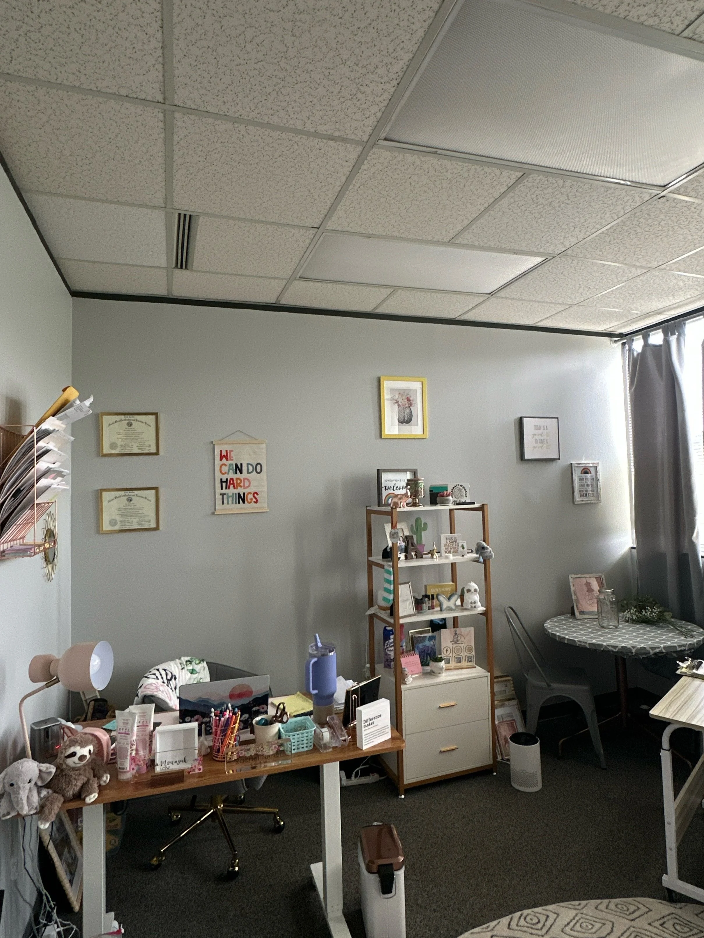 Office space with a desk, shelves, and a small round table, decorated with plants and framed pictures, and various office supplies and plush toys.