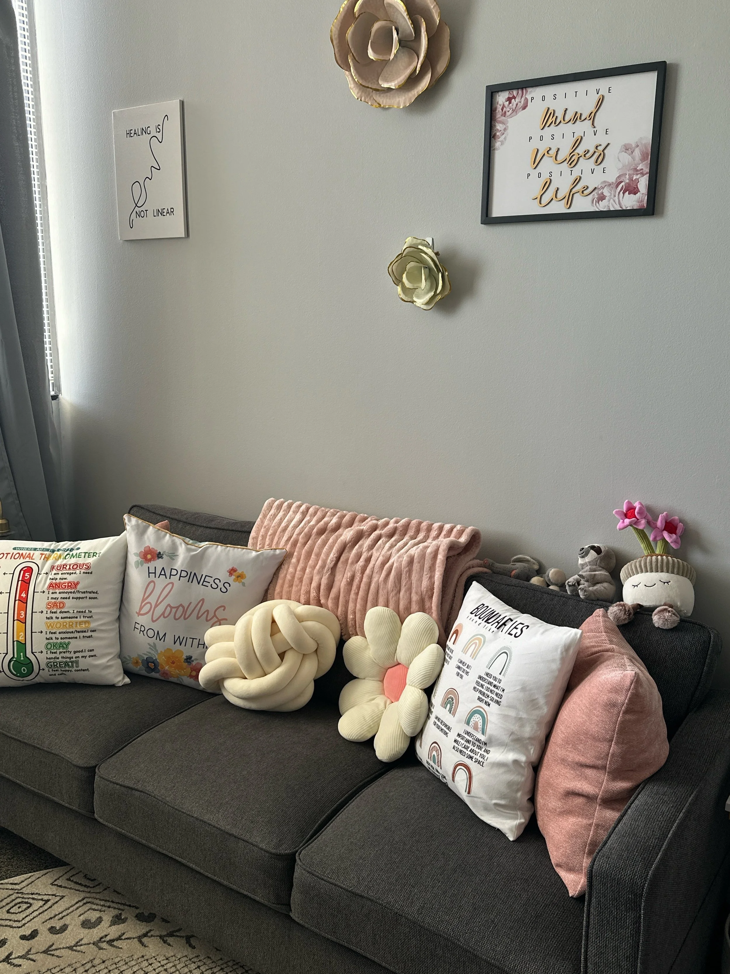 Living room with gray sofa decorated with colorful pillows, plush toys, and pink throw blanket. Wall art includes motivational quotes and floral pictures.