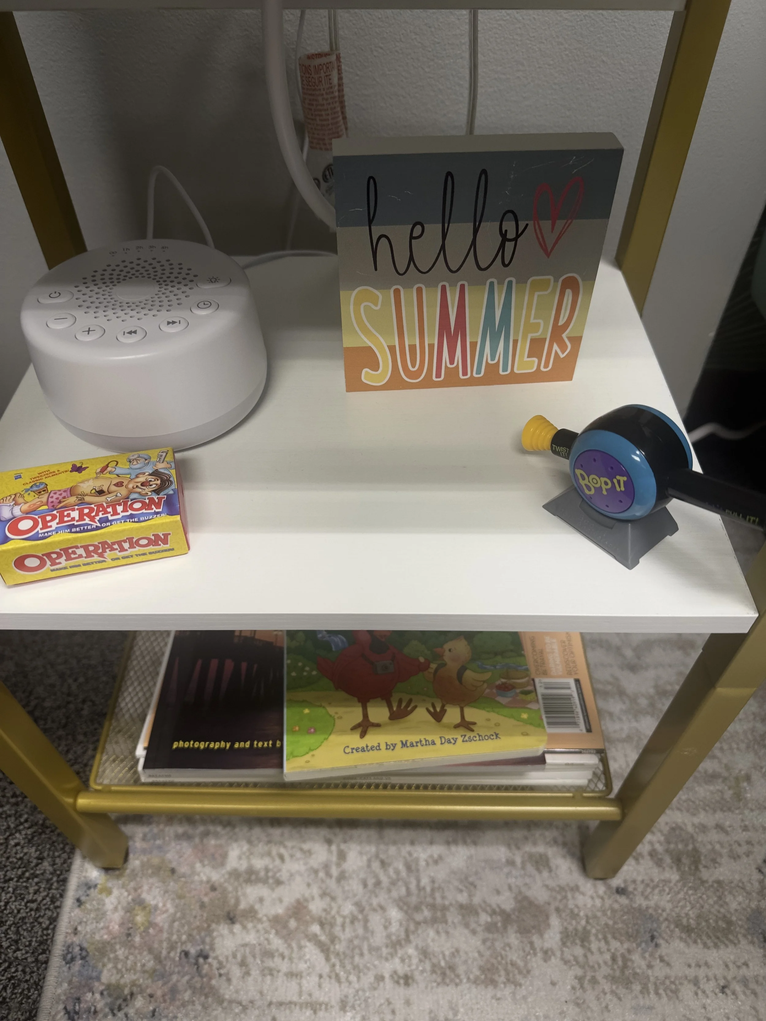 White shelf with a colorful sign that says "hello summer" and a heart, a white radio or speaker, a box of Operation game, a handheld game called Bop It, and books on the lower shelf.