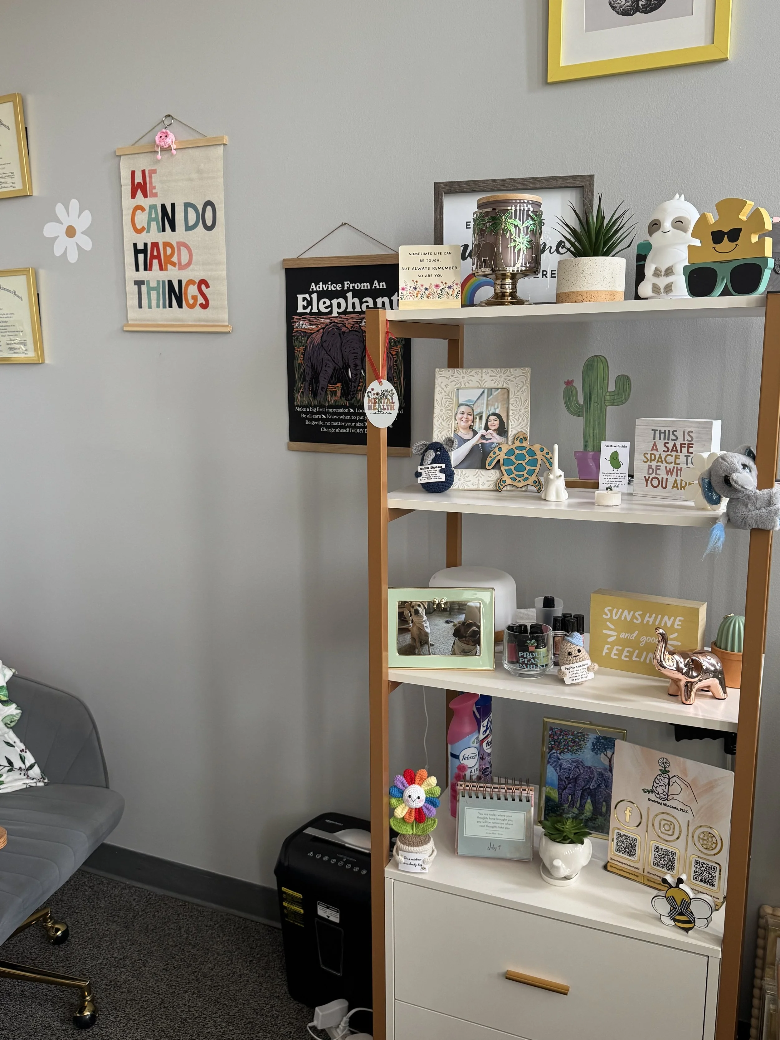 A white shelf with decorations, framed photos, small plants, stuffed animals, and colorful signs, set against a gray wall with hanging art and inspirational quotes.