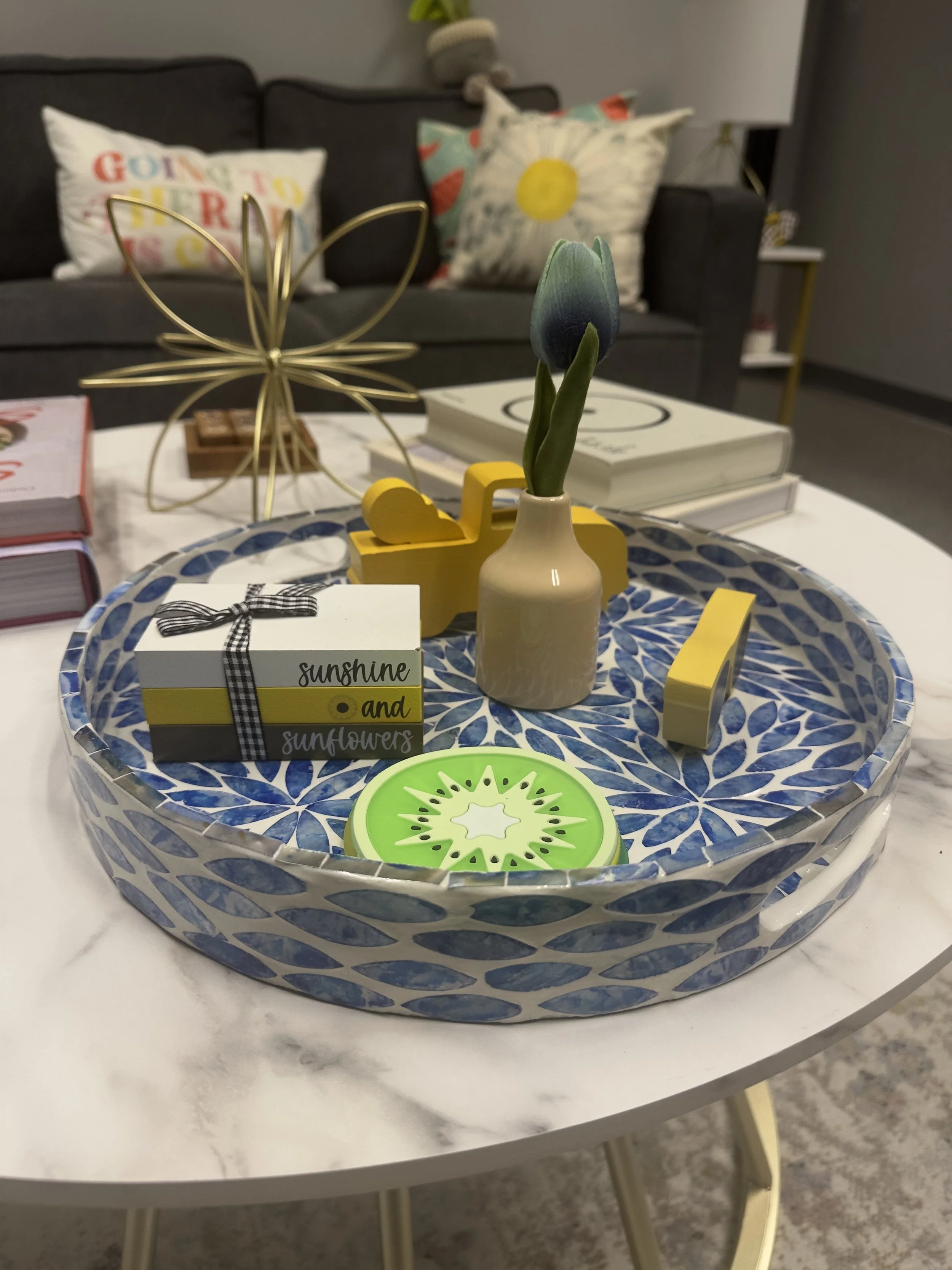 Decorative tray on a white marble table with a small vase containing a tulip, a green circular candle, and a gift box with a black ribbon. In the background, there are books on the tray, a gold wire butterfly sculpture, and a gray sofa with colorful cushions.