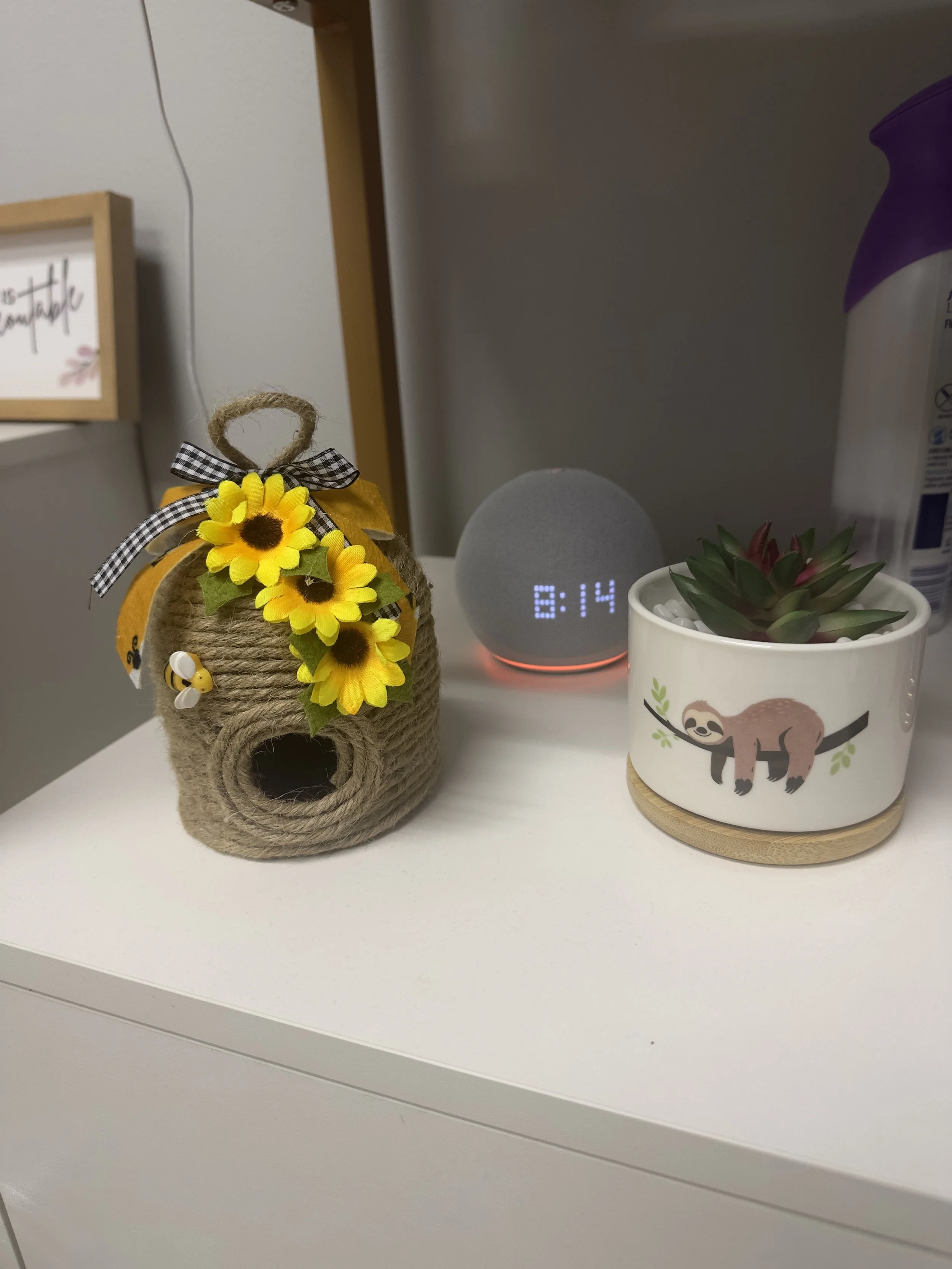 Decorative sunflower-shaped birdhouse with a black and white checkered bow and small bee embellishment, next to a white mug with a sloth illustration, a digital clock displaying 8:14, and a potted succulent plant, on a white surface.