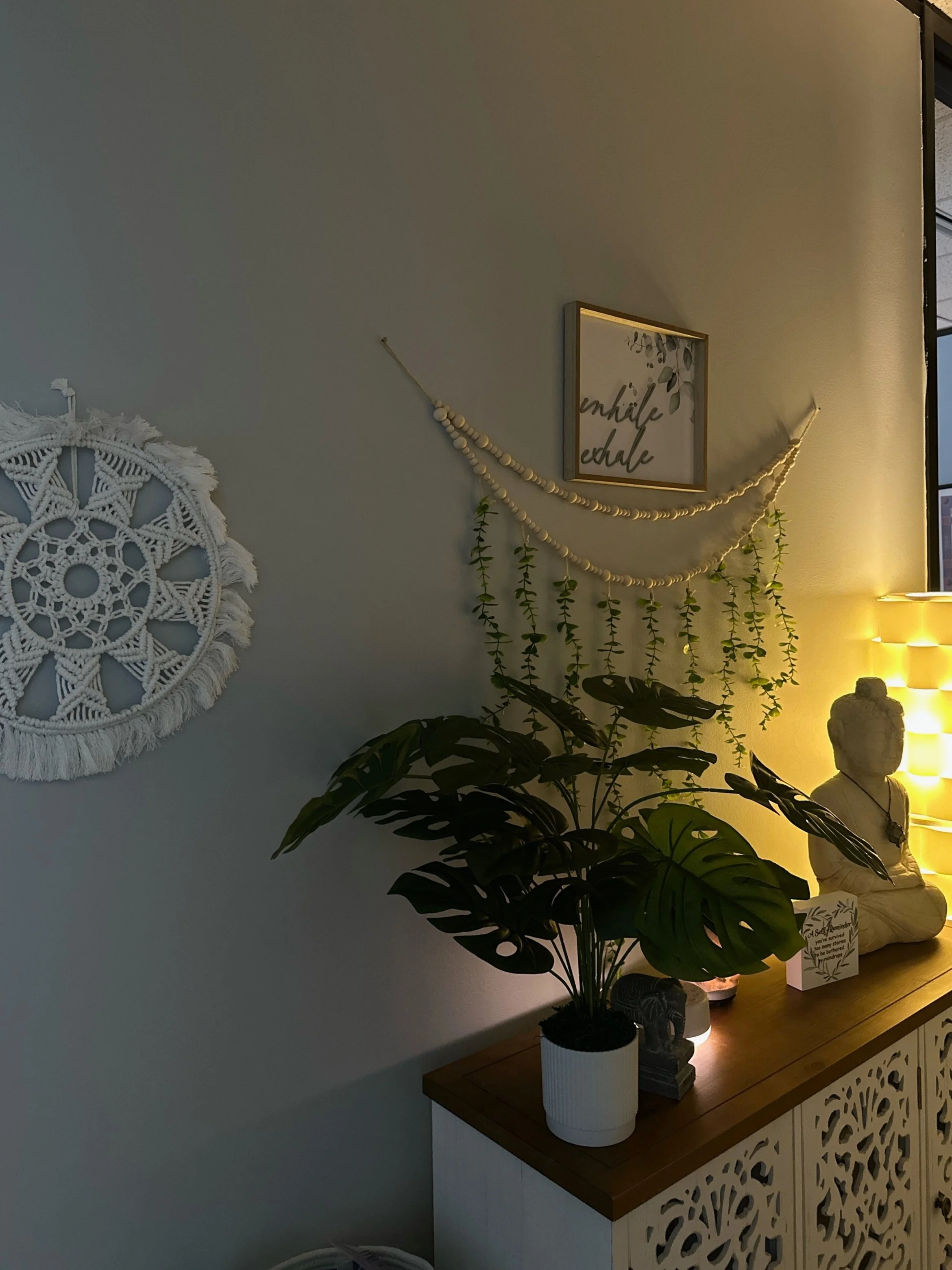 Decorative interior space featuring a white crochet wall hanging, framed art with the text "inhale exhale" and green hanging plants, a wooden sideboard with a large monstera plant in a white pot, a Buddha statue, and warm lit lamps.