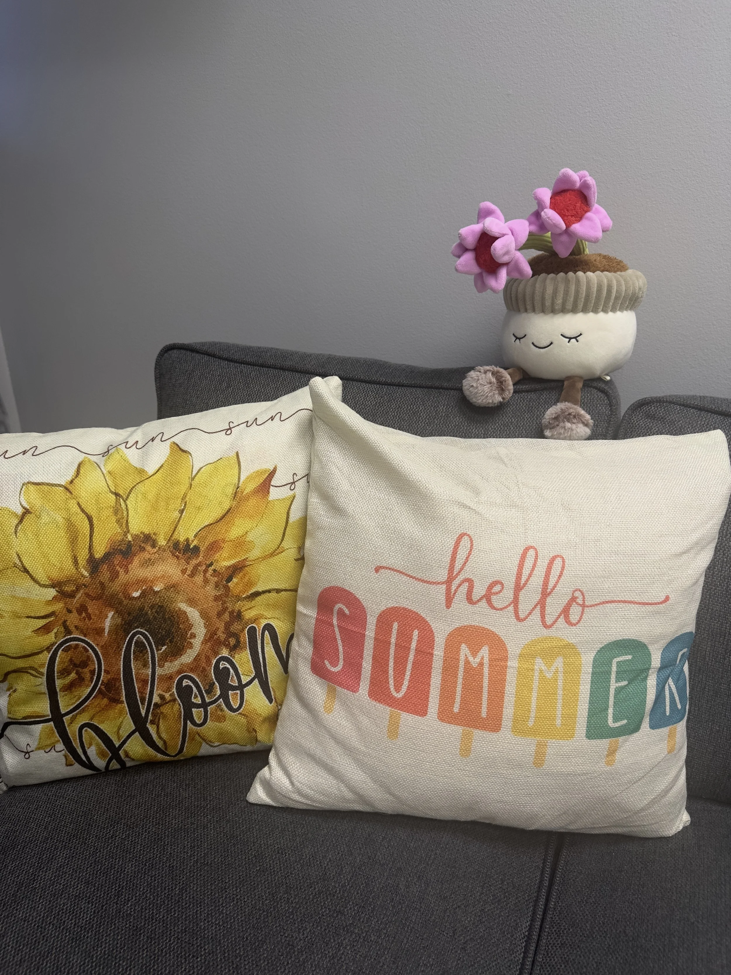 Decorative pillow with sunflower design and the word 'bloom' on a gray couch, and a cream-colored pillow with the words 'hello summer' in colorful letters, with a plush planter with pink flowers on top of the couch.