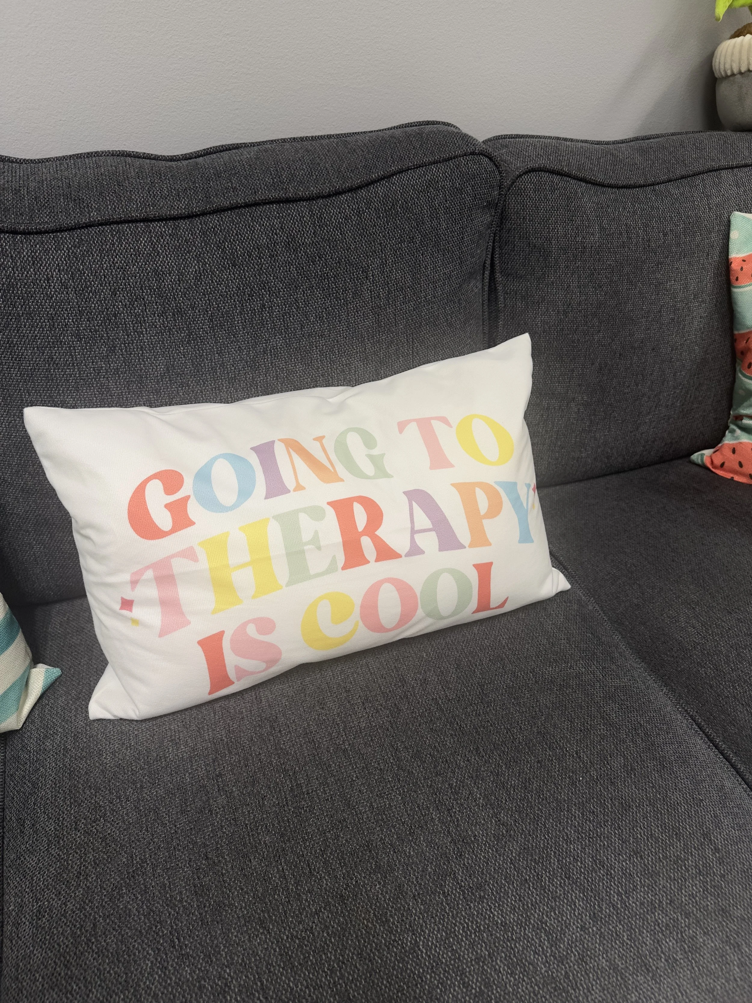 Decorative pillow on dark gray sofa with colorful text that reads 'Going to therapy is cool'