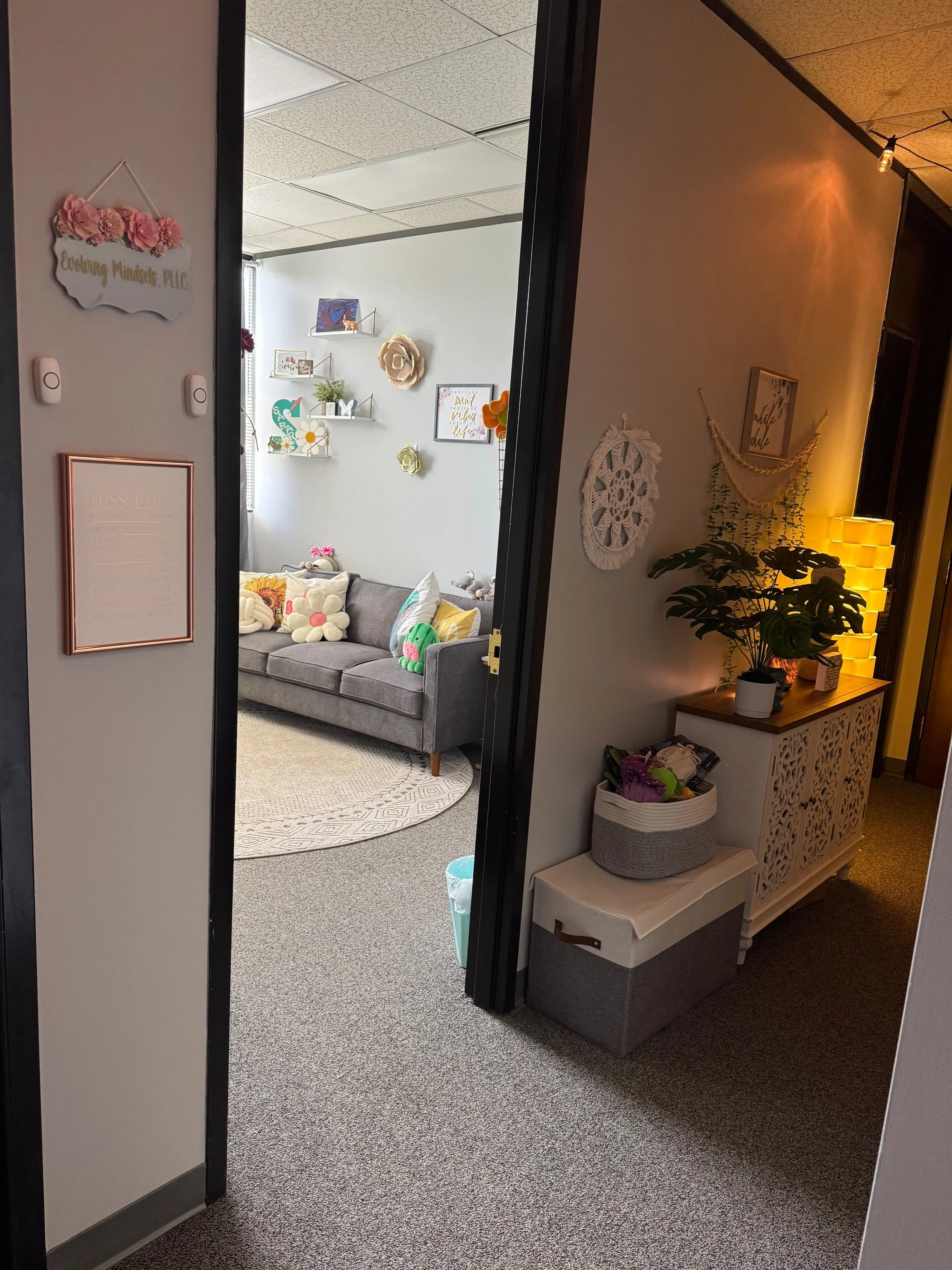 View into a cozy office room decorated with wall art, shelves, plush pillows on a gray couch, and a large green plant, with a seating area in the hallway featuring a white cabinet with intricate cut-out designs and a small bench with a basket on top.