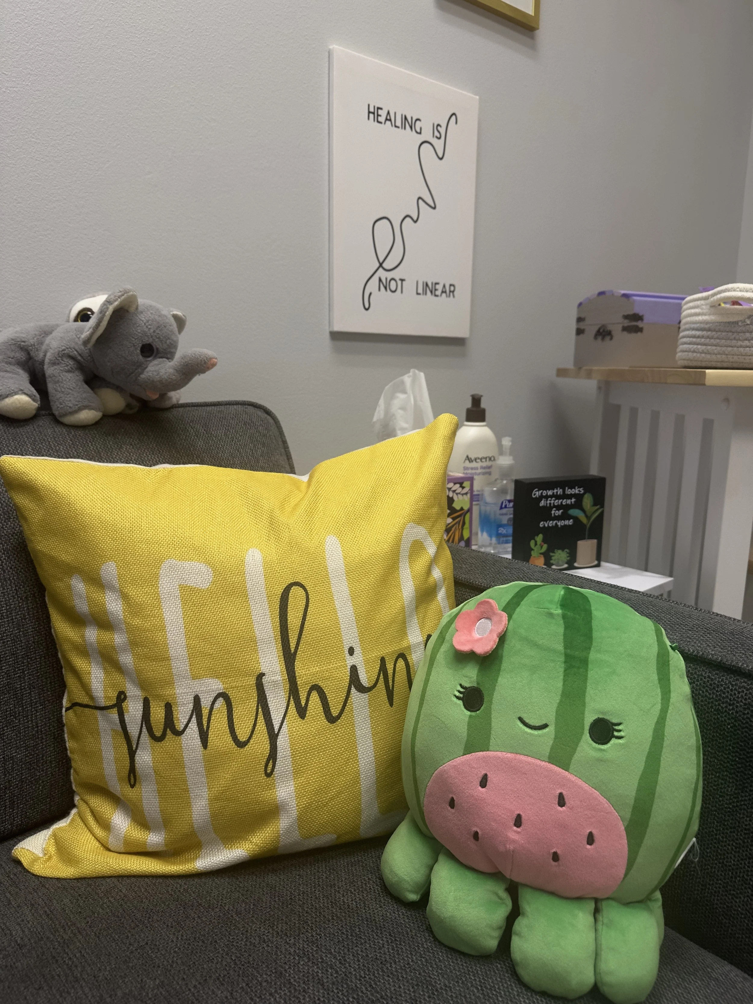 Decorative plush watermelon-shaped pillow with a pink flower on it and a yellow pillow with white and black text on a dark grey couch. A stuffed grey elephant toy is on the back of the couch. On the wall, a white framed quote reads, "Healing is not linear," with a line drawing of a snake.