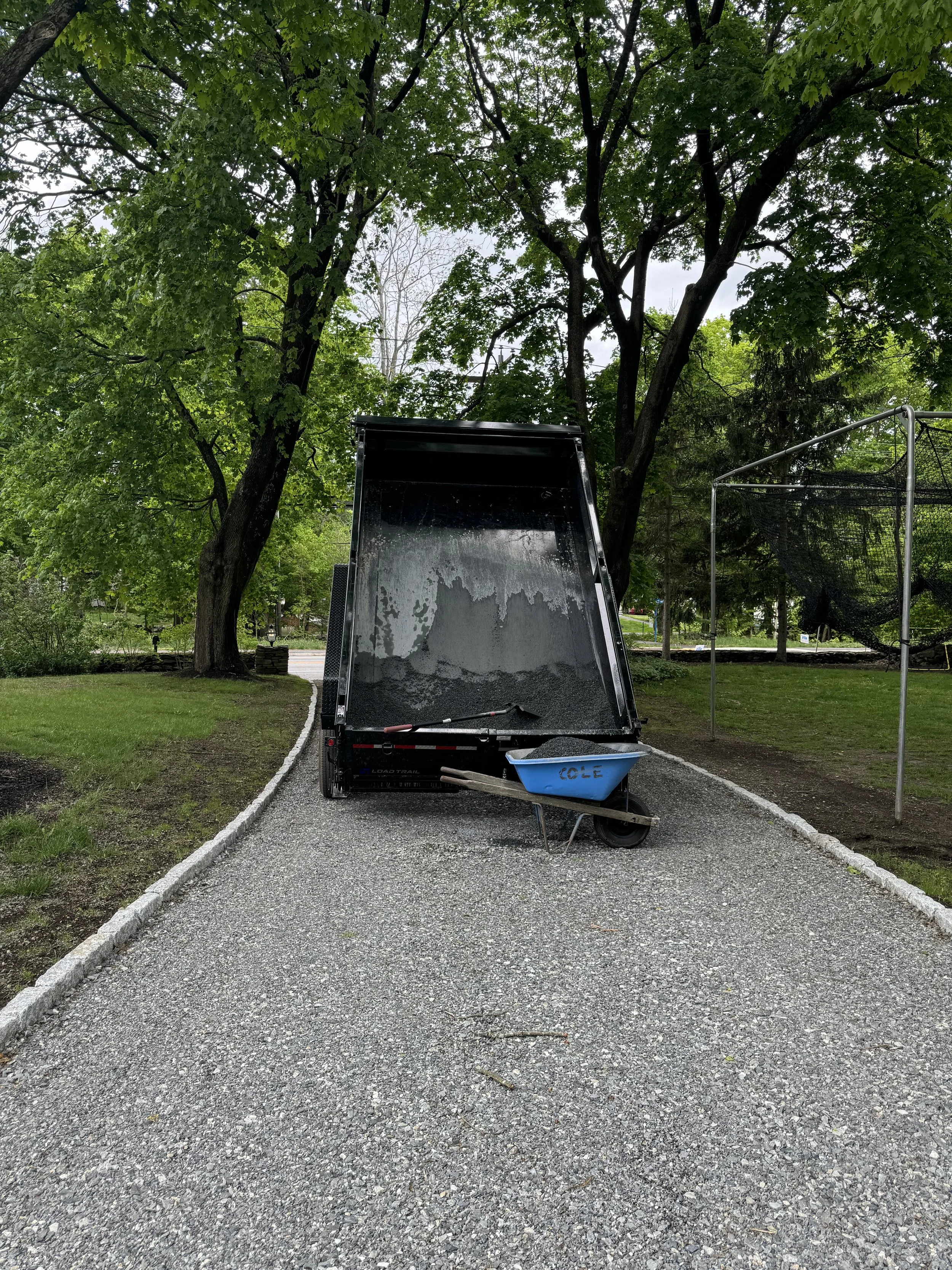Gravel driveway beside a white house with a landscaped garden featuring green plants and a young tree with pink flowers.