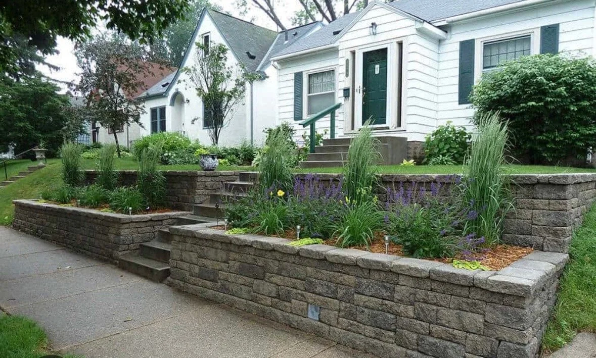 Side of a house with white siding, bordered by a stone walkway and a garden area with sparse plants and soil.