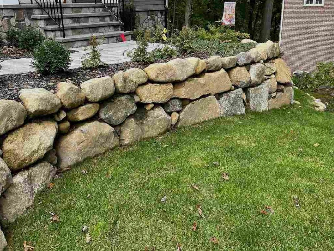 Construction site with partially built stone wall along house, soil and gravel, tools including a wheelbarrow, brush, and rubber mallet.