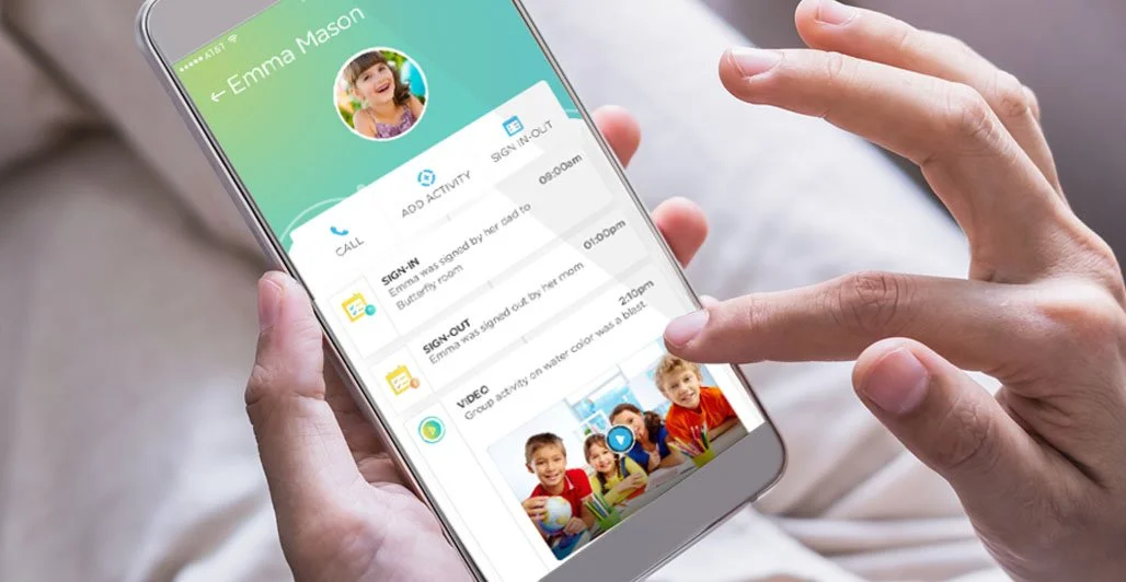 A person holding a smartphone displaying a child monitoring app with a profile picture of a smiling girl named Emma Mason. The app shows call logs, sign-in/out activity, and a video thumbnail of children in a classroom.