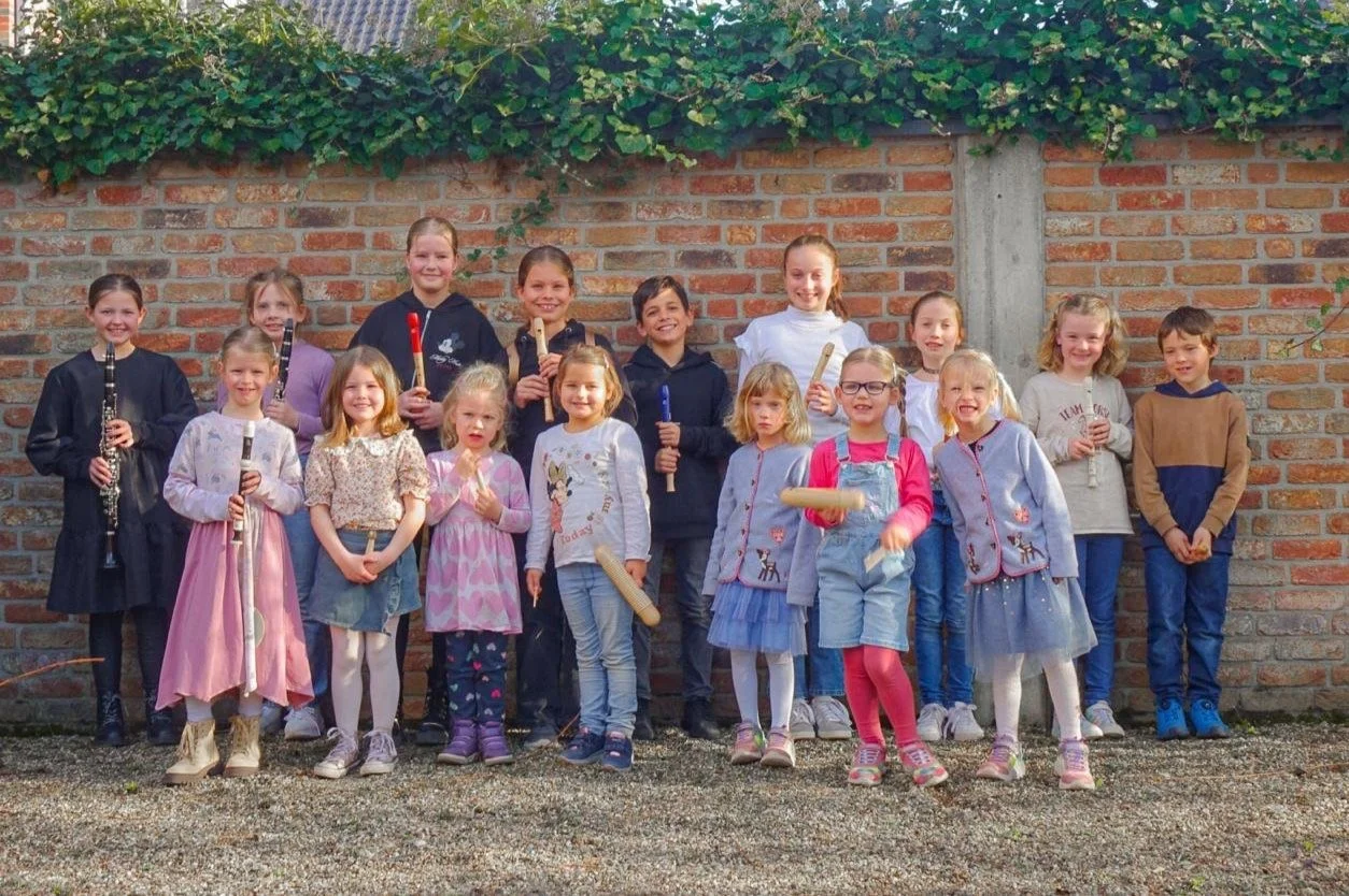 Gruppe von Kindern vor einer Backsteinwand, viele halten Musikinstrumente in der Hand. Sie lächeln in die Kamera und tragen bunte, legere Kleidung. Über ihnen wächst Efeu entlang der Mauer.