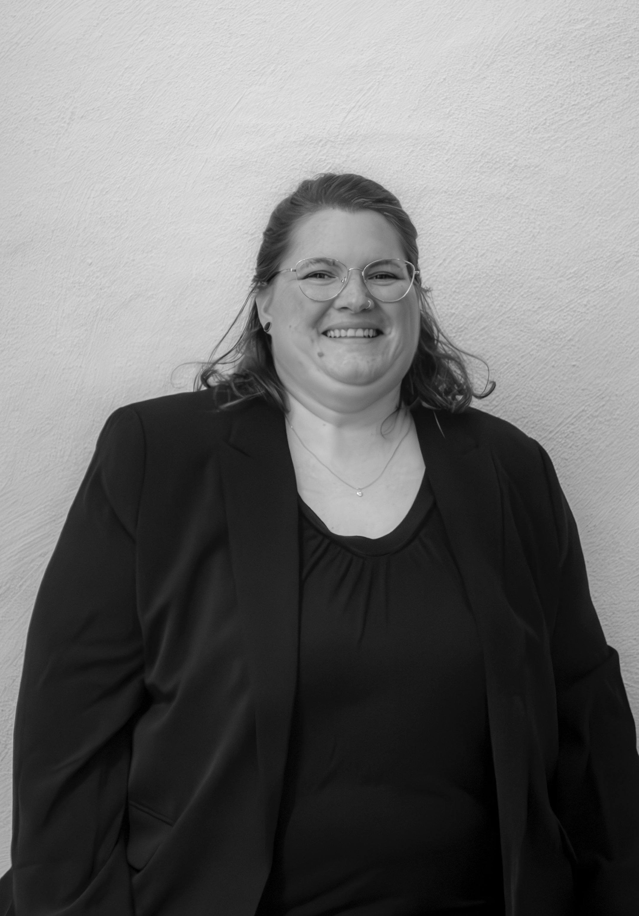 Schwarz-weiß Foto einer Person in einem Blazer, die lächelt, vor einer hellen Wand.